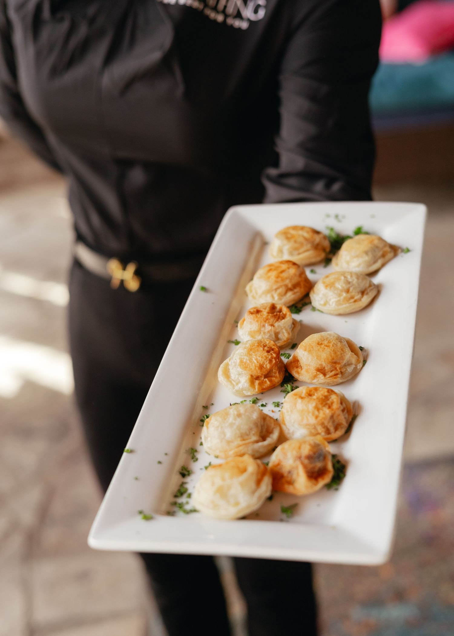 Server holding a white rectangular platter of golden-brown dumplings garnished with fresh herbs at a catered event.
