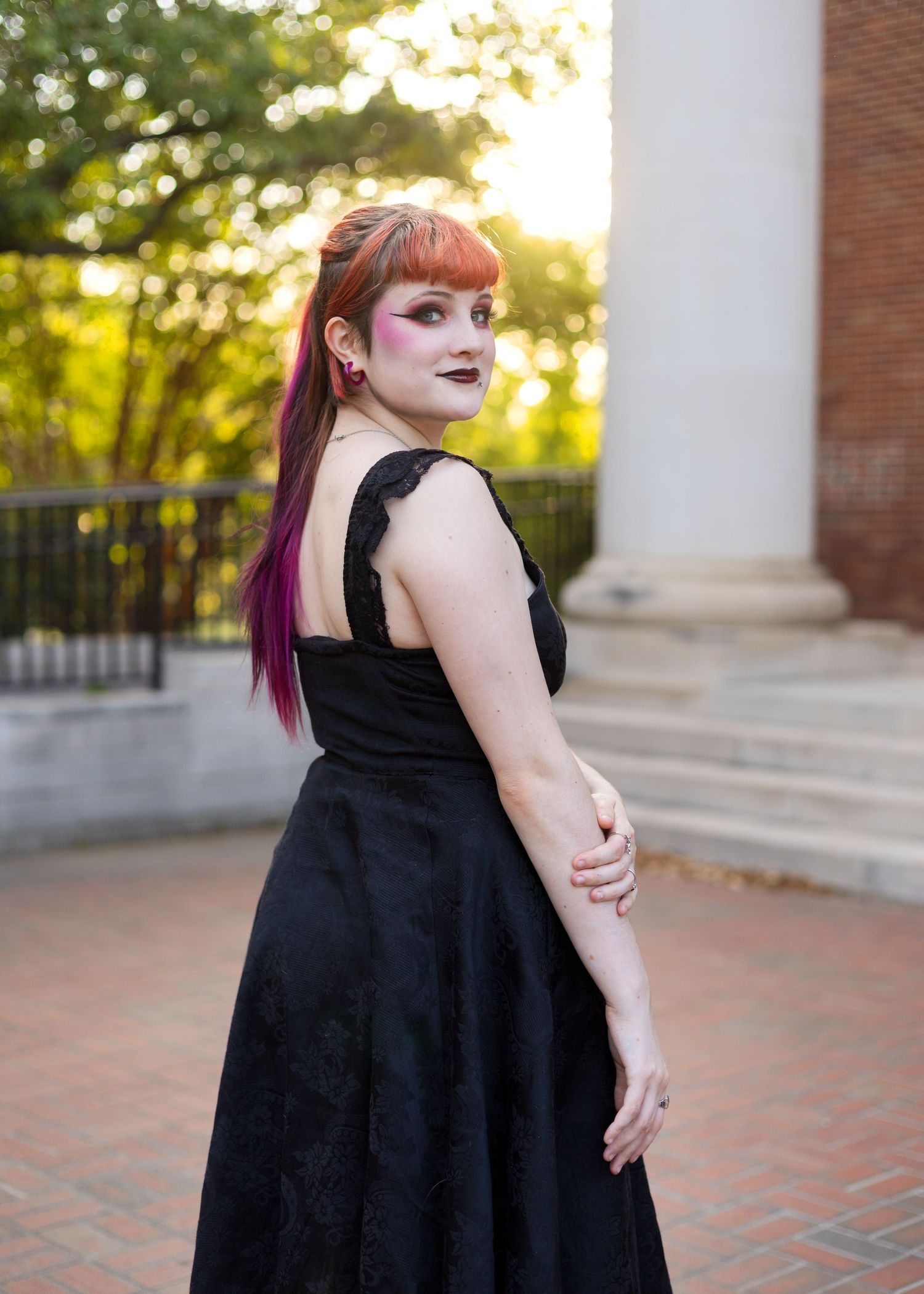 Woman in black backless gown with red-purple hair poses outdoors on brick plaza at golden hour.