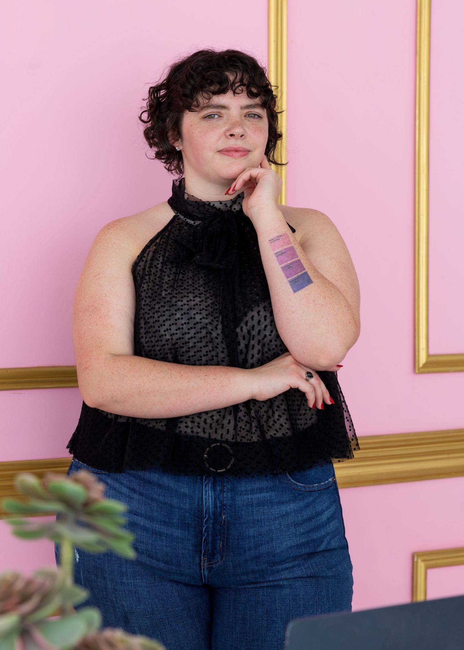 A person in a black sleeveless top and jeans poses against a pink wall with gold trim molding.
