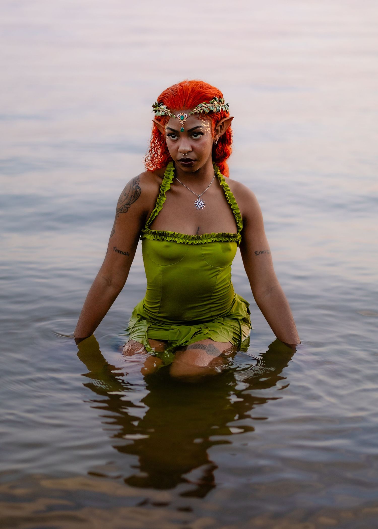 Figure in bright green dress and floral crown poses dramatically in water at sunset.