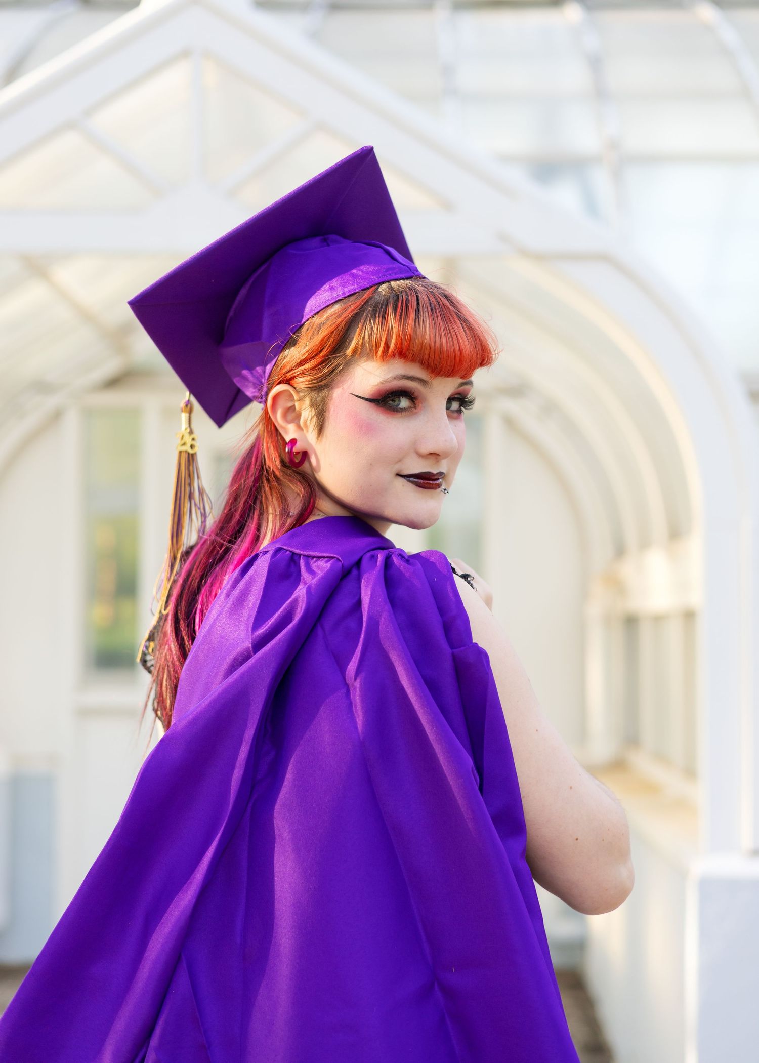 Young woman in purple graduation cap and gown with red hair poses outdoors, looking back over shoulder.
