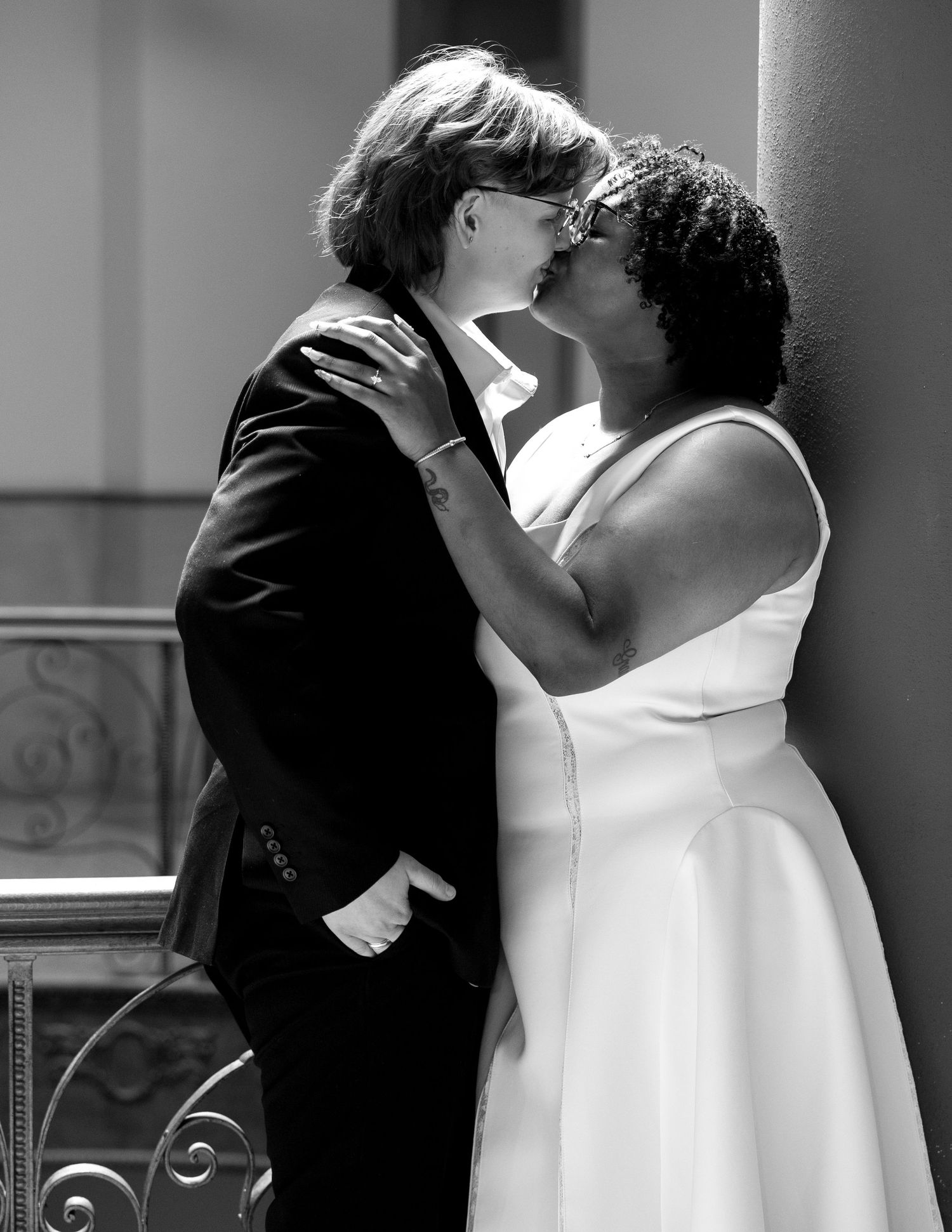 Black and white photo of two women in wedding attire sharing a kiss, one in a suit and one in a white dress.