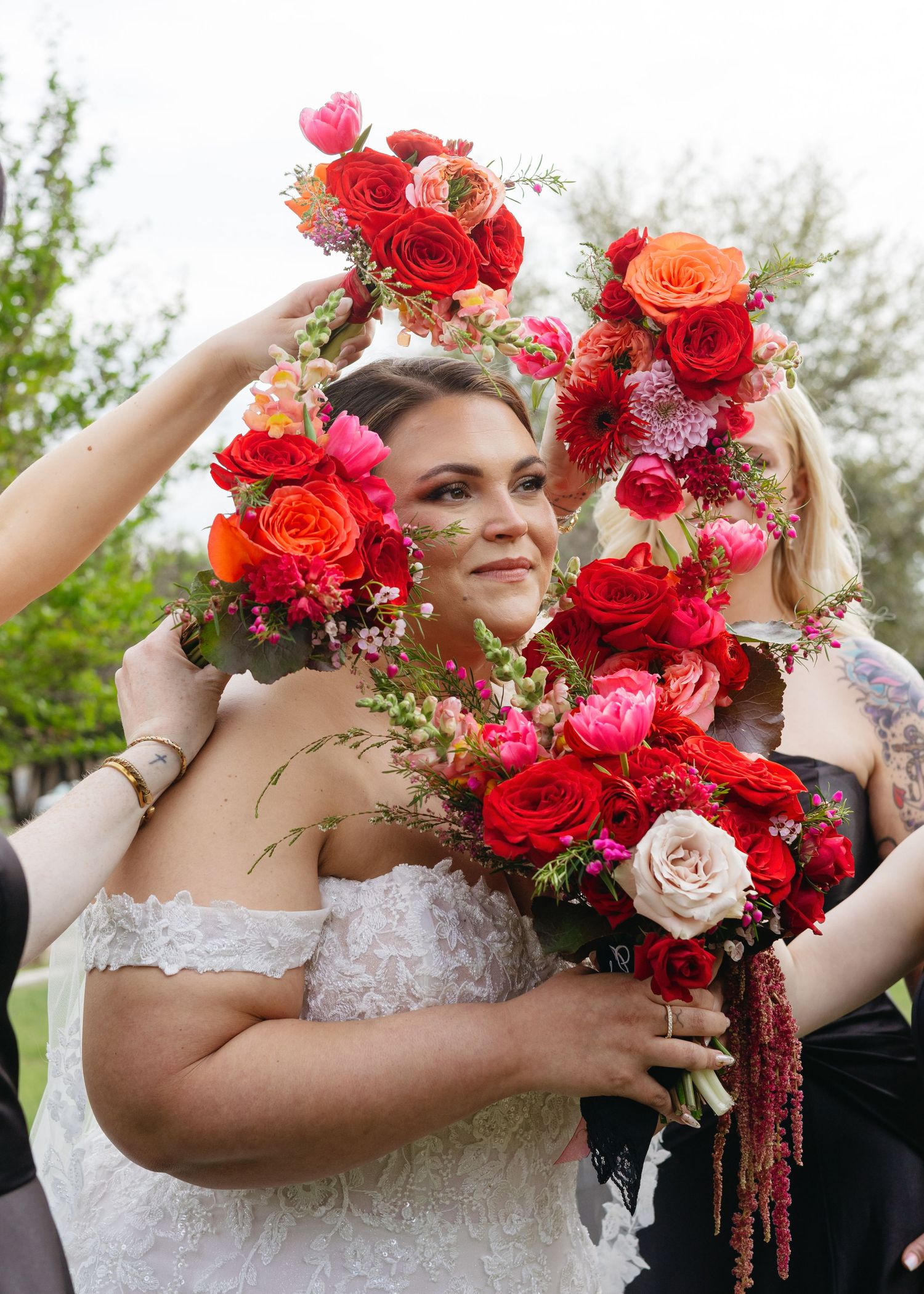 Bride in lace gown surrounded by vibrant red, pink, and orange wedding bouquets held by smiling bridesmaids.