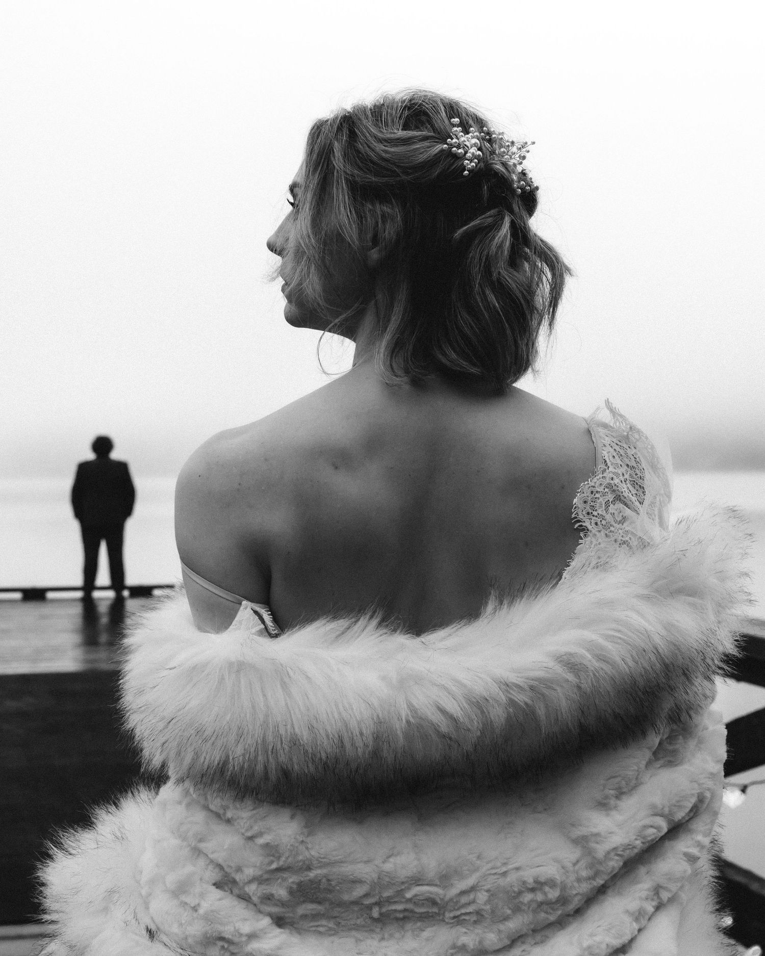 Bride in lace gown and fur wrap views groom by misty waterfront, black and white wedding photo.
