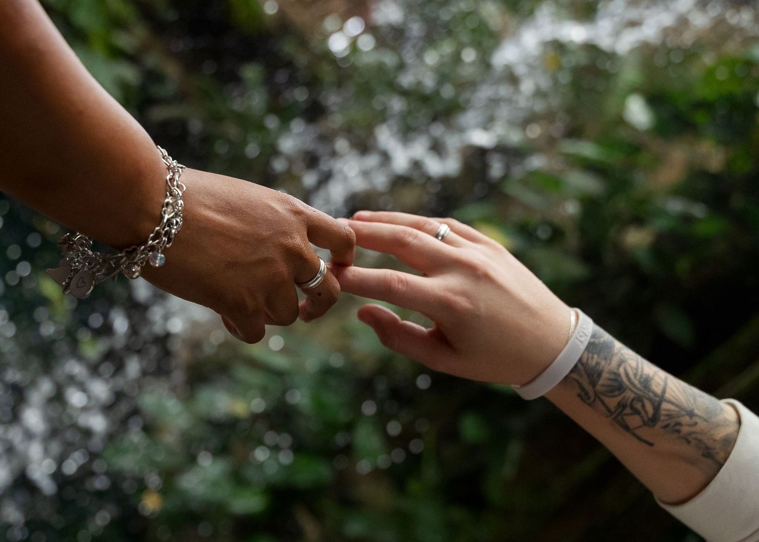Two hands gently touching, one with a charm bracelet and one with a floral tattoo and diamond ring, with greenery behind.