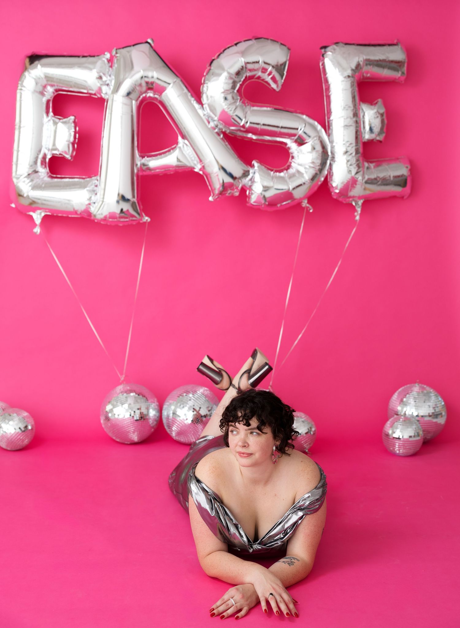 Silver balloon letters float against bright pink backdrop while someone poses playfully below.