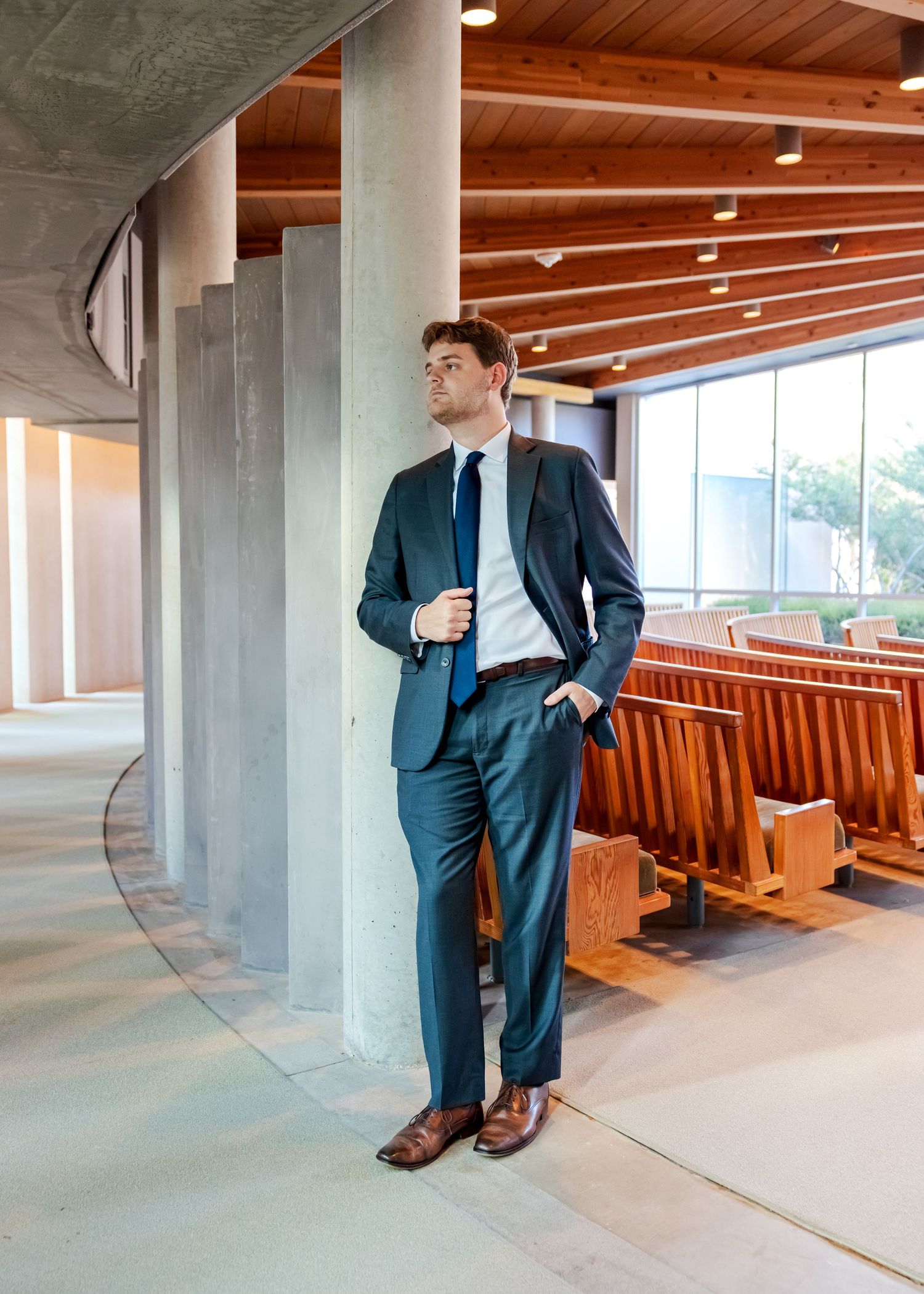 Man in teal suit with blue tie stands confidently in modern wood-ceiling chapel with pew seating.