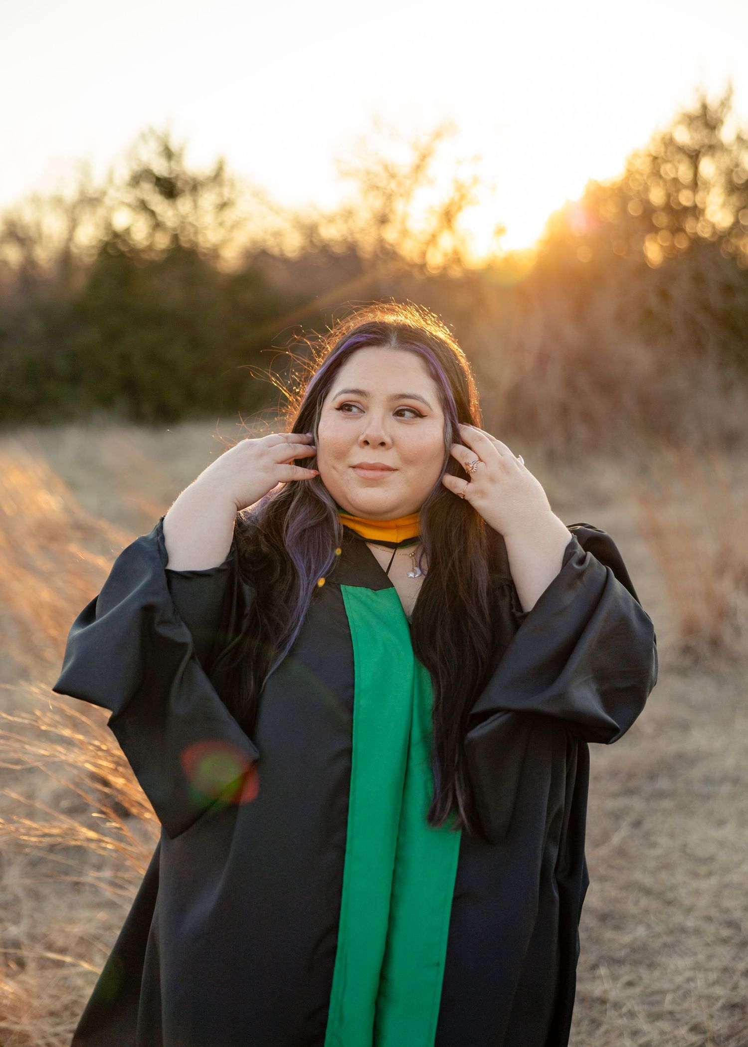 A person in a black coat and green scarf poses in a field during golden hour sunset.