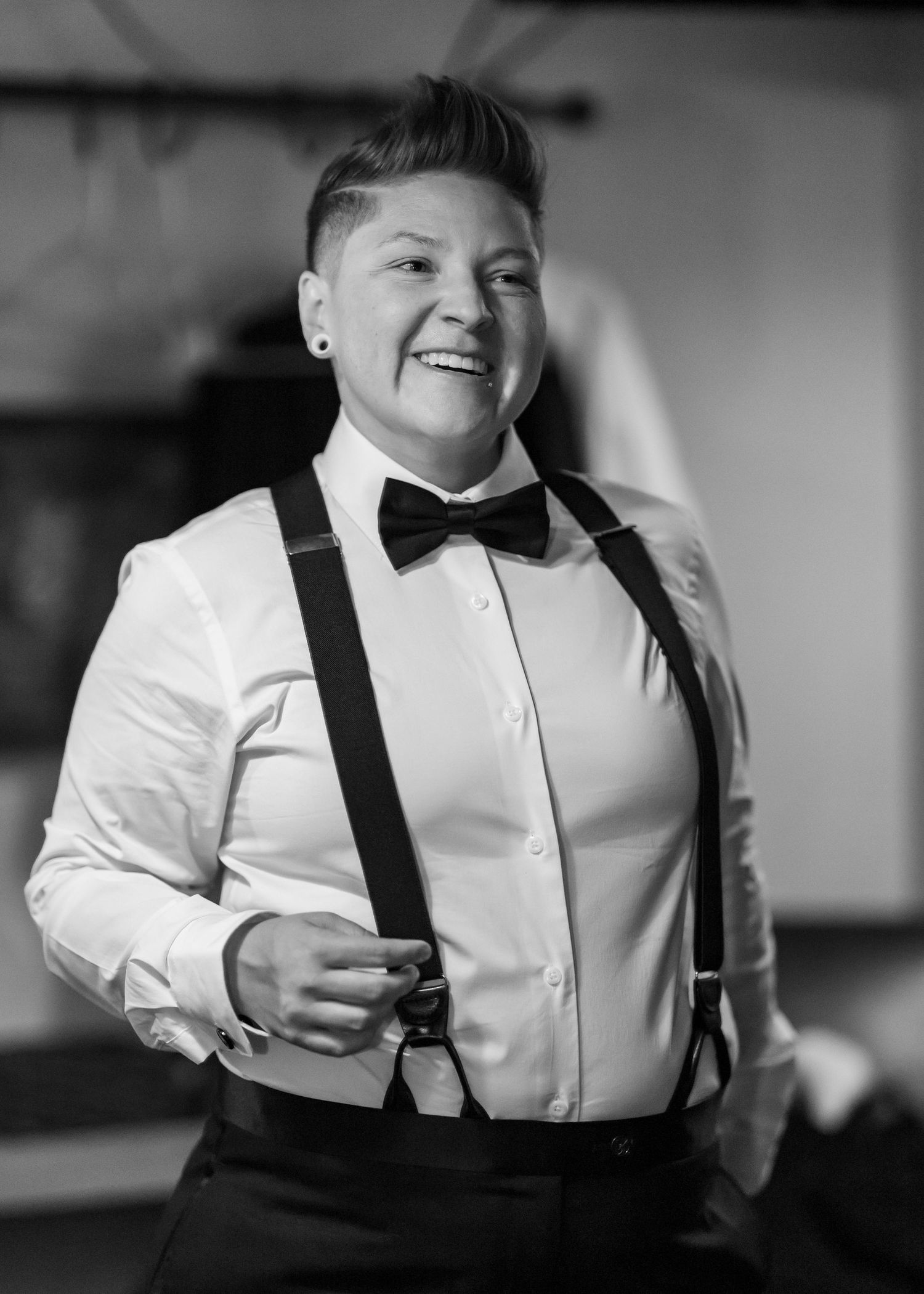 Person in white dress shirt, suspenders, and bow tie smiling joyfully at a formal event.