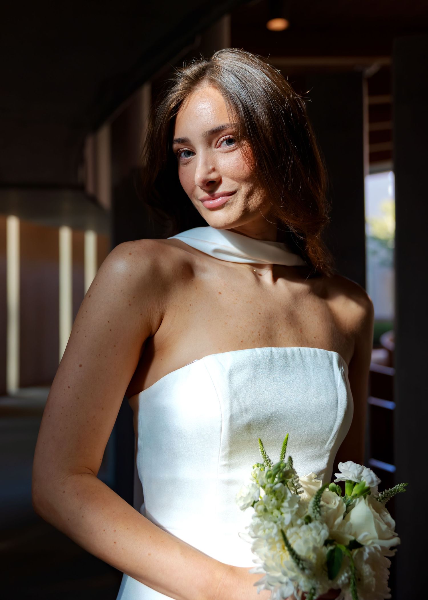 Elegant bride in strapless white gown holding white floral bouquet in soft natural light.