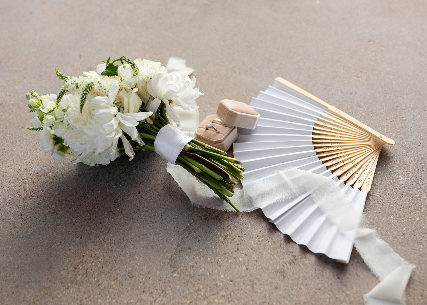 White peony bouquet with ribbon beside an elegant white paper hand fan on a gray surface.