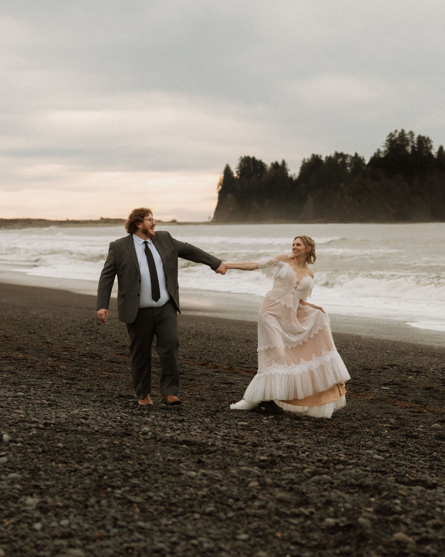 A couple in wedding attire holds hands on a dark sandy beach with ocean waves and evergreen trees in the background.
