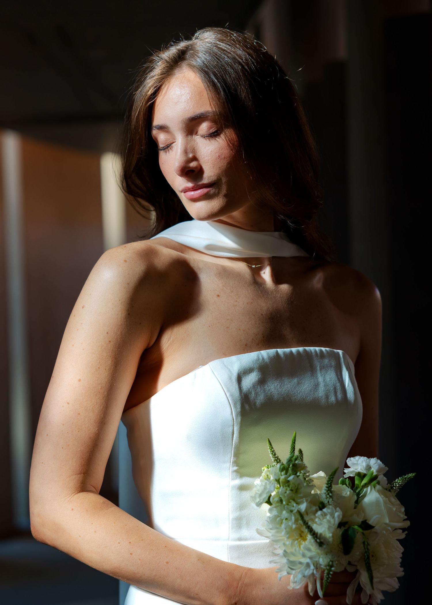 Bride in white strapless gown with halter detail holds bouquet of white flowers in soft natural light.