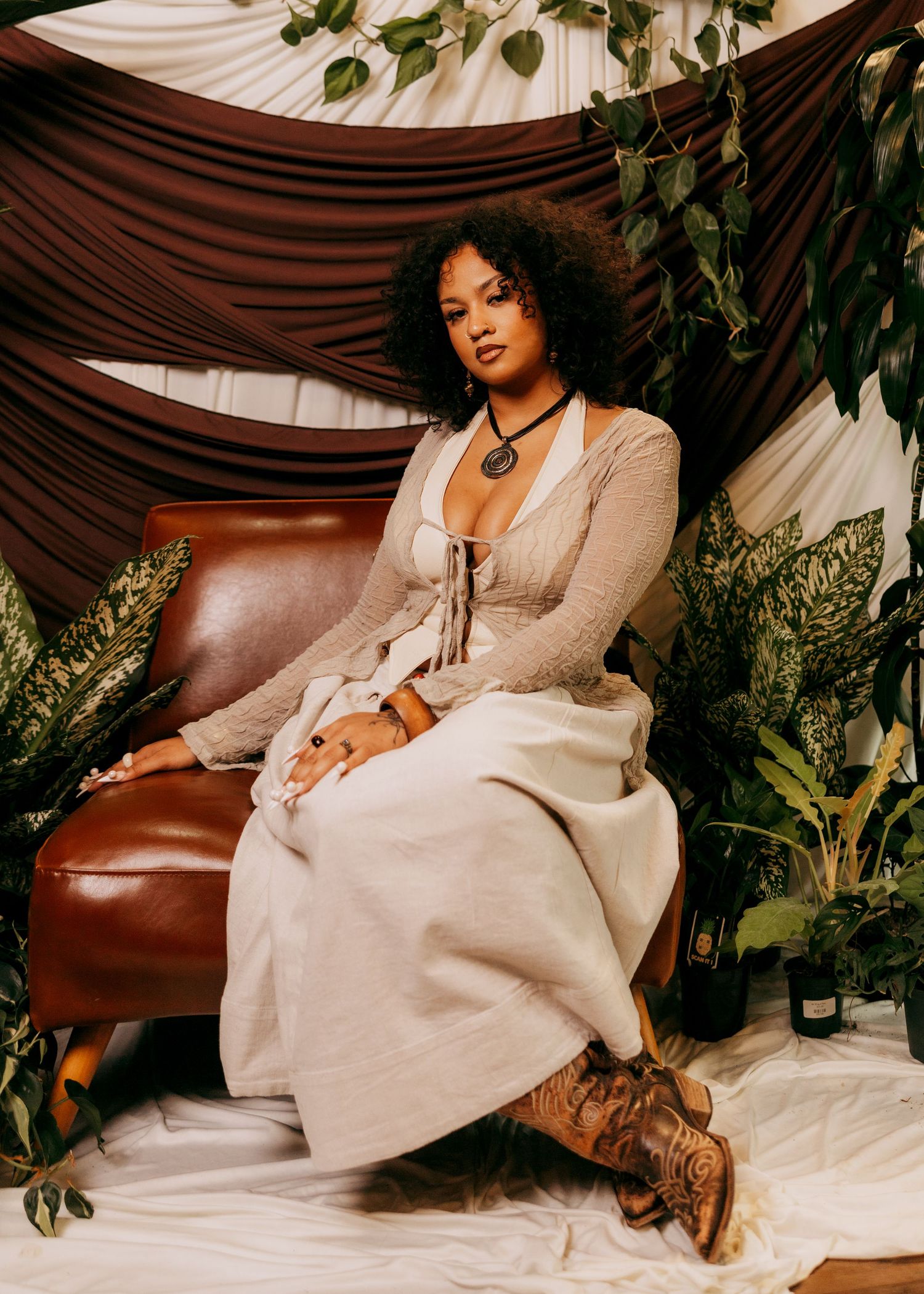 Person in beige dress lounging on brown leather couch surrounded by plants.