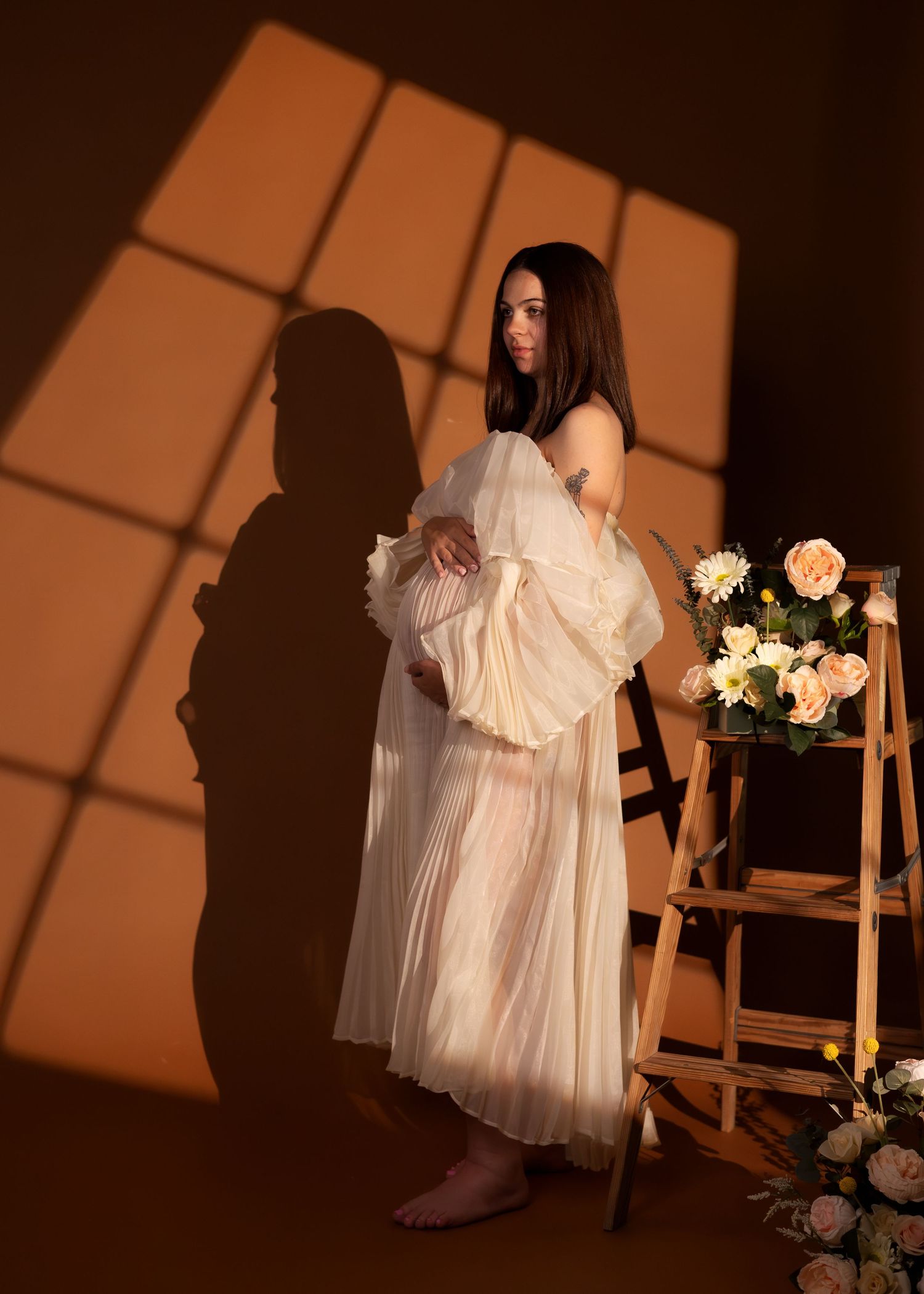 Pregnant woman in sheer white gown poses by floral arrangement on wooden ladder, dramatic shadow light on brown wall.