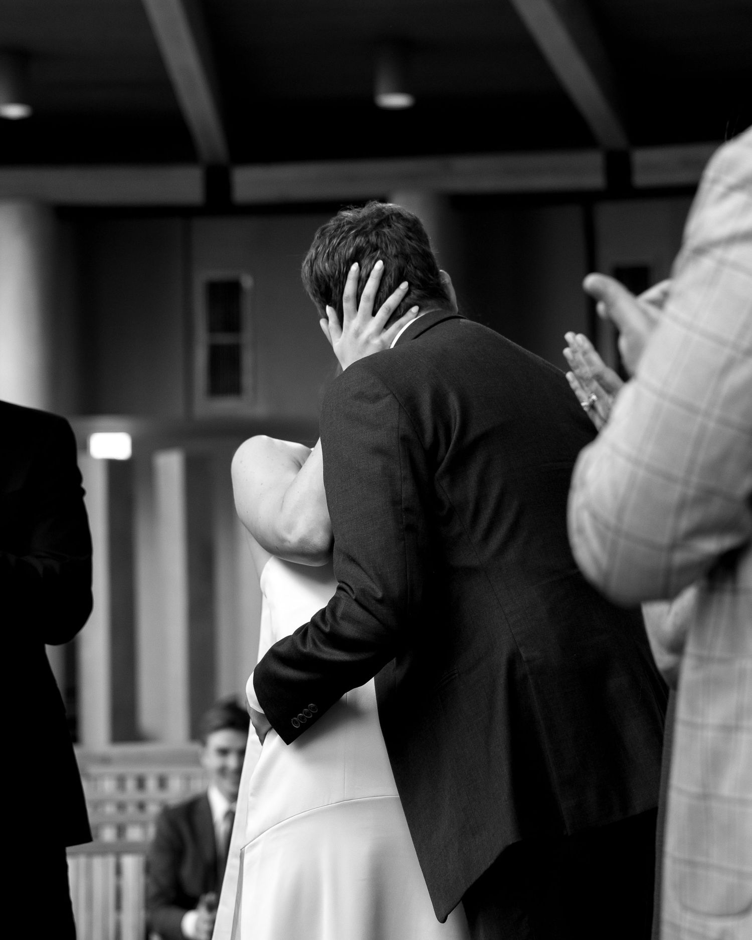 Black and white photo of a couple sharing their first kiss at a wedding ceremony while guests applaud.