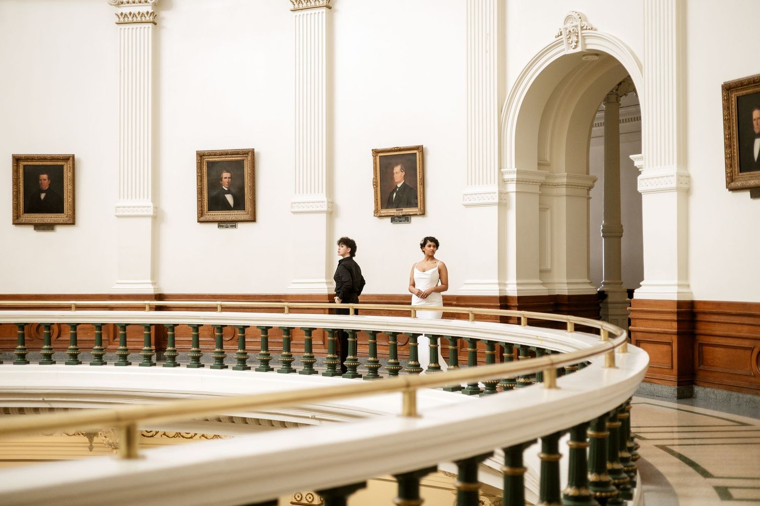Bride and groom share a romantic moment on a curved balcony in an elegant white hall adorned with portrait paintings.