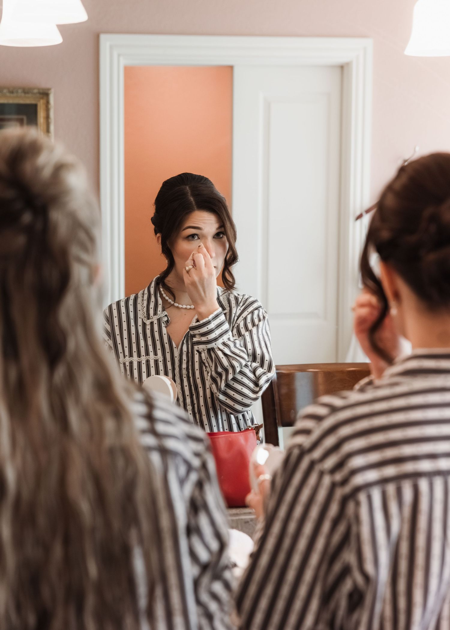 A bride in striped pajamas touches her face emotionally while reflected in a mirror surrounded by bridesmaids.