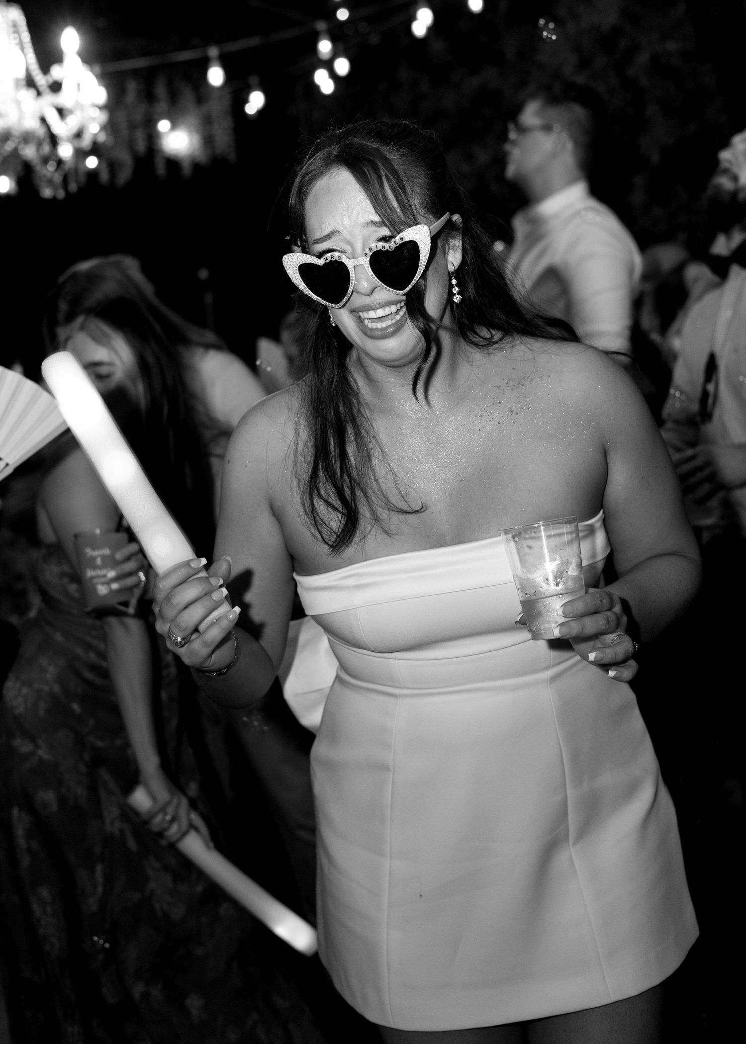 Woman in white dress and heart-shaped sunglasses dancing at a party, holding a glow stick and drink.