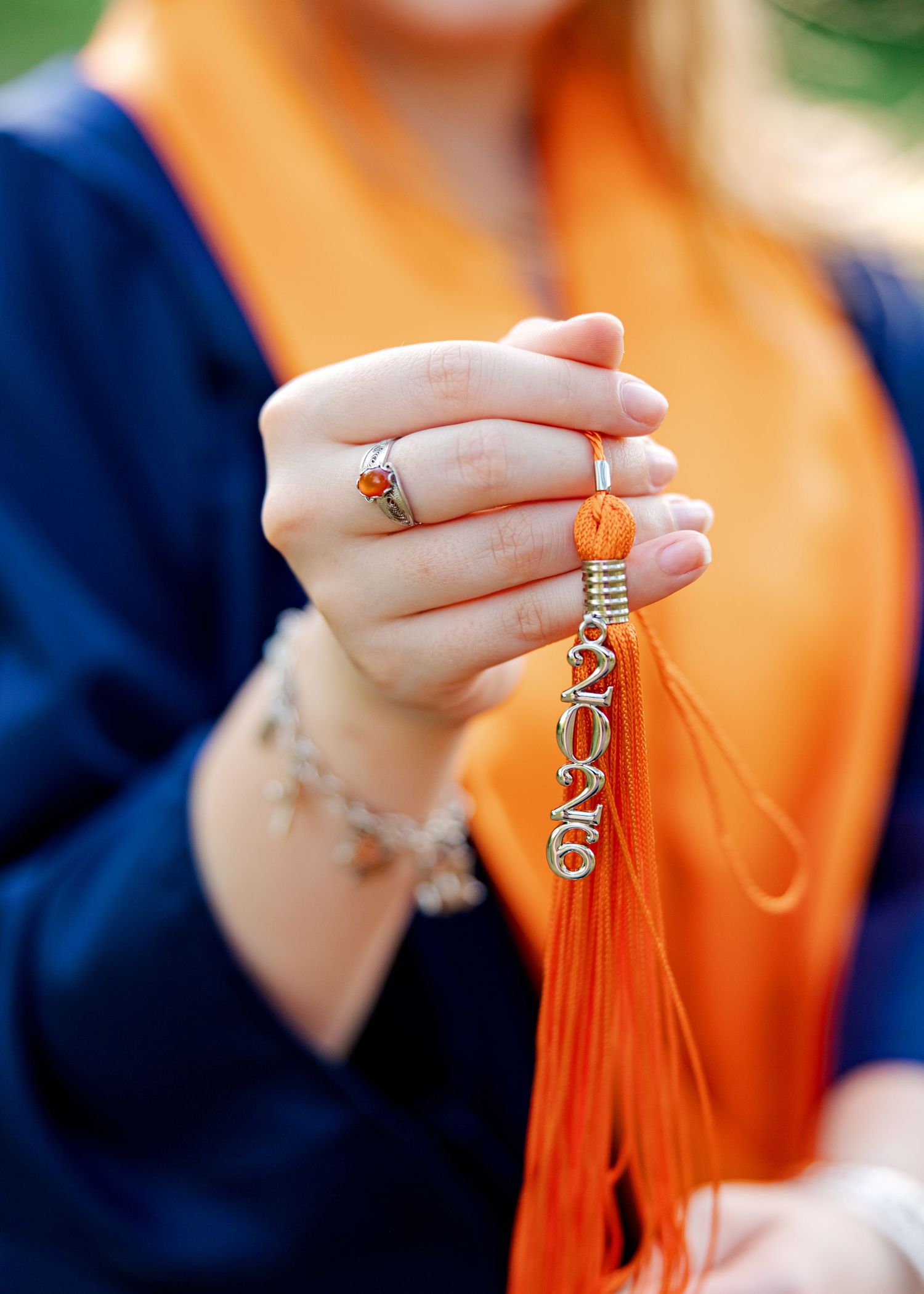 Close-up of hands holding an orange graduation tassel with 2026 charm, wearing a ring and bracelet.
