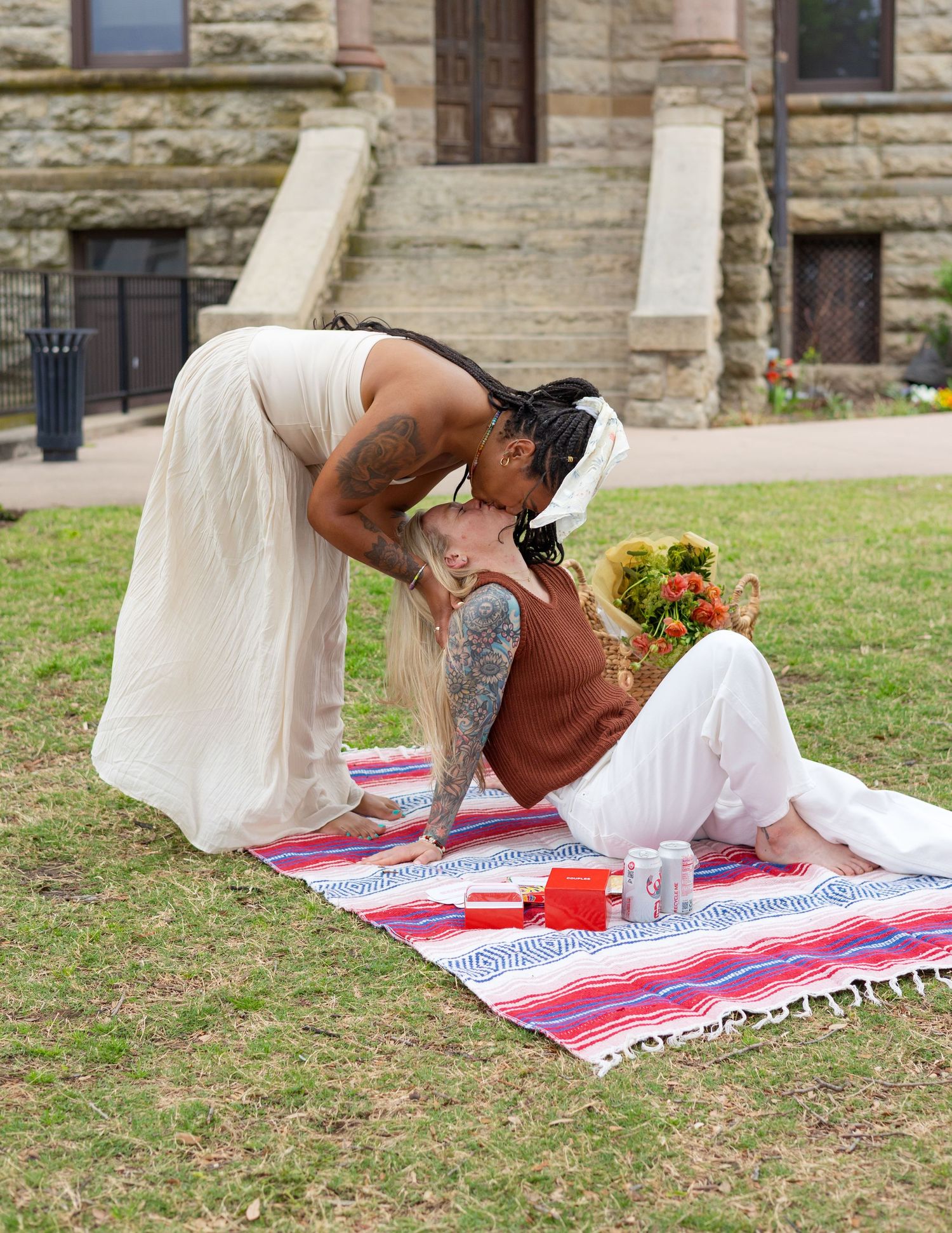 Two brides share a romantic kiss on a colorful blanket outdoors during a wedding ceremony.