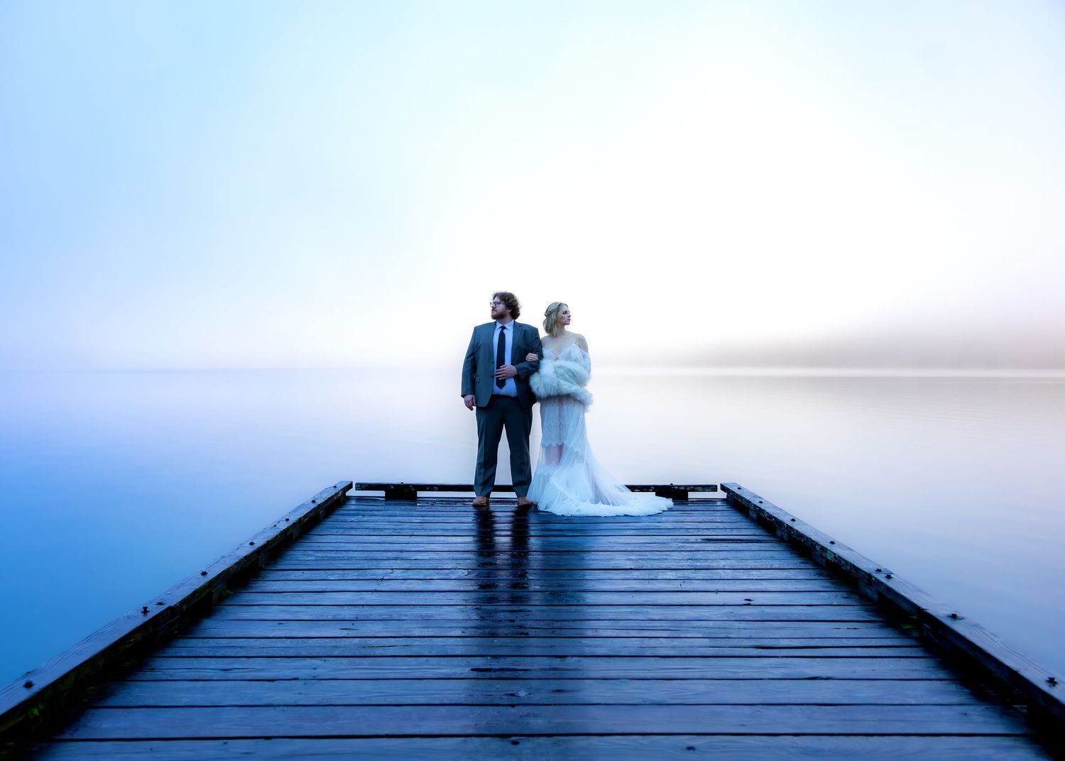 Couple stands at the end of a blue wooden dock surrounded by fog.