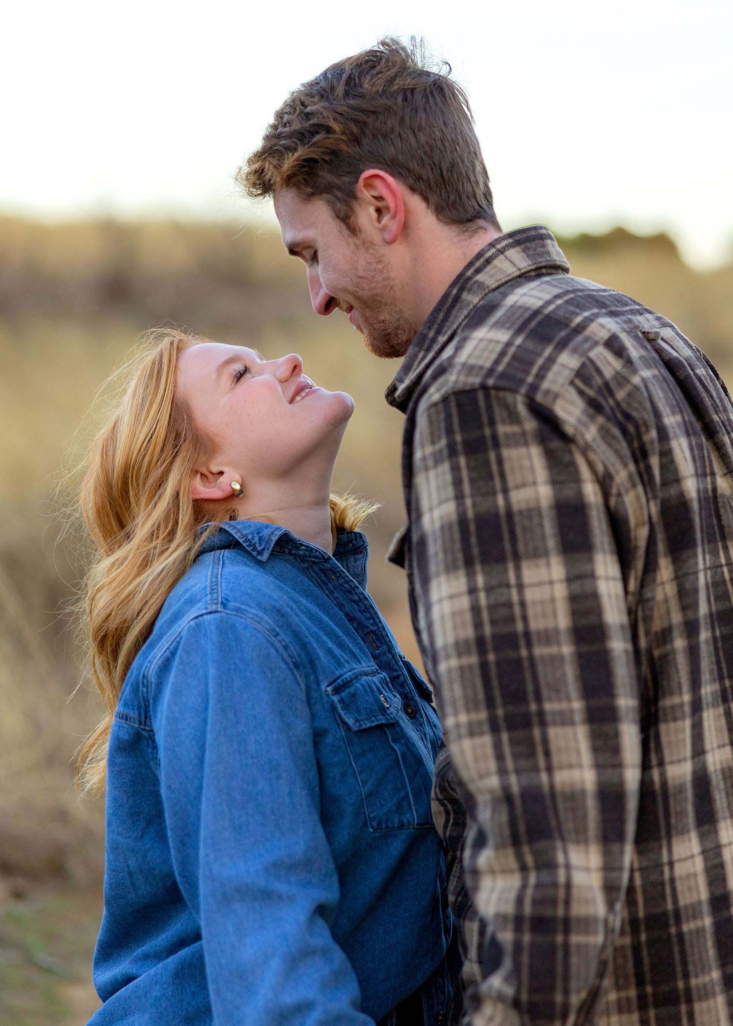Couple in denim and plaid share tender moment in outdoor field setting.