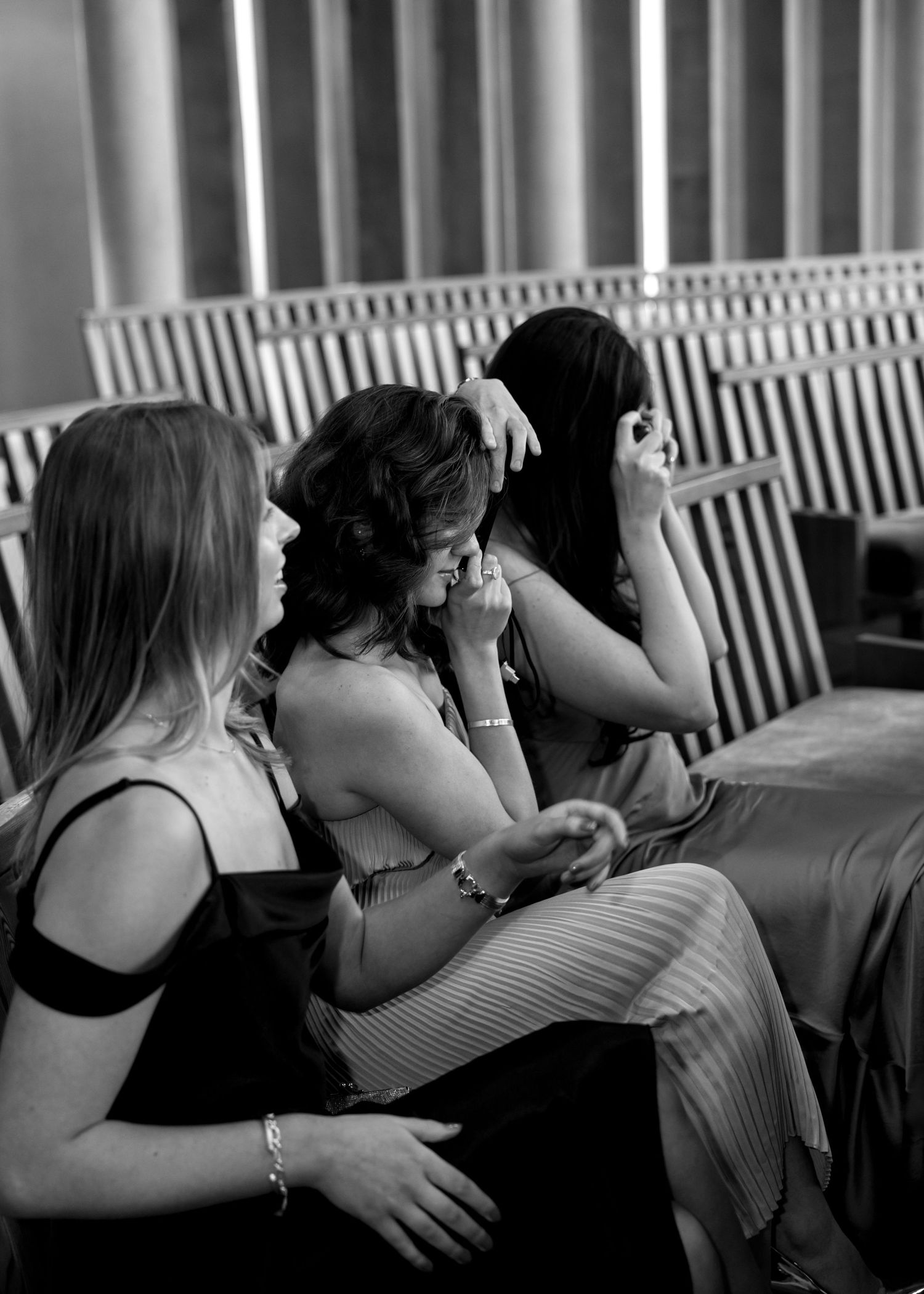 Three women share an emotional moment seated together in a venue during a wedding ceremony in black and white.