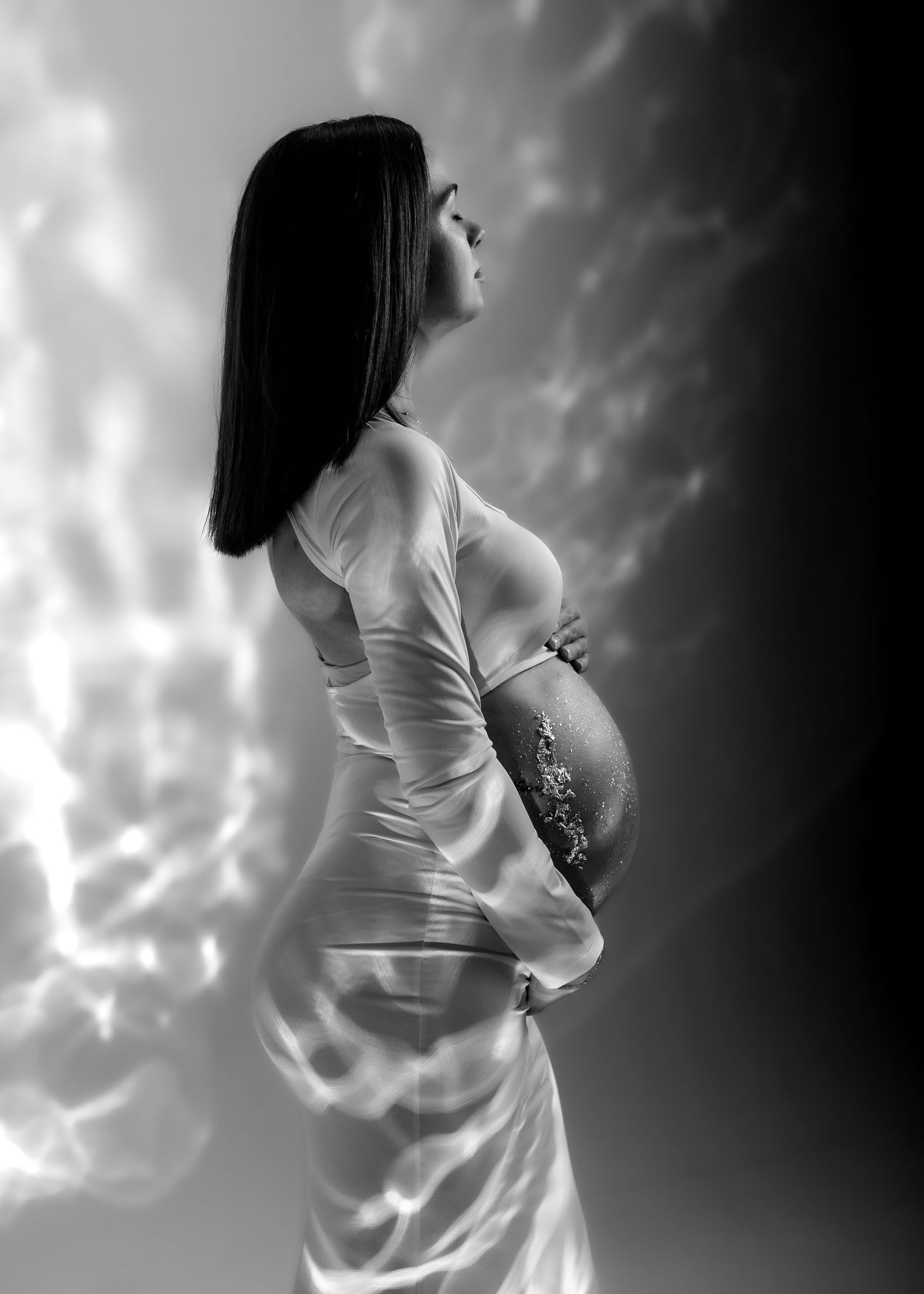 Black and white maternity photo of pregnant woman in white dress holding belly against dramatic cloudy sky backdrop.