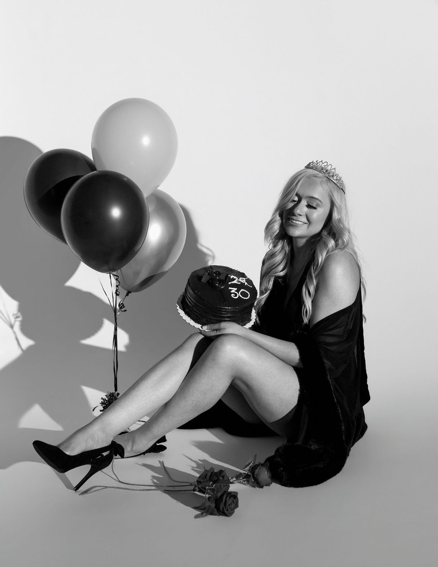 Woman in black dress and tiara sits on floor holding 30th birthday cake with balloons and rose in black and white photo.