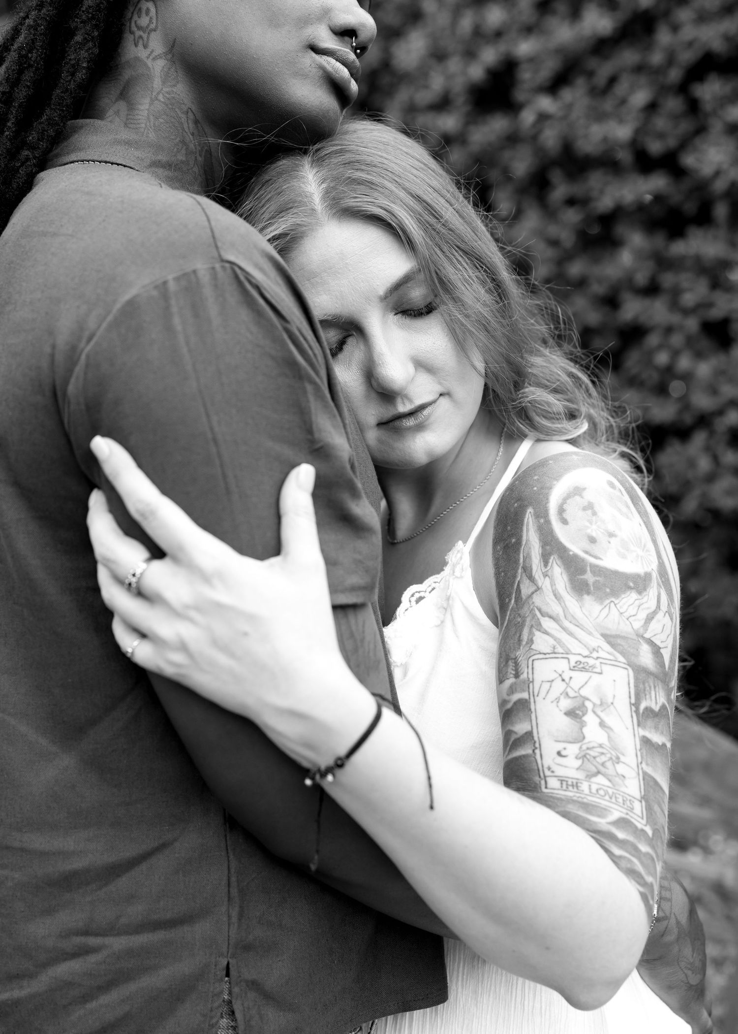 Black and white photo of a couple embracing, woman with tattoos resting her head on a mans chest.