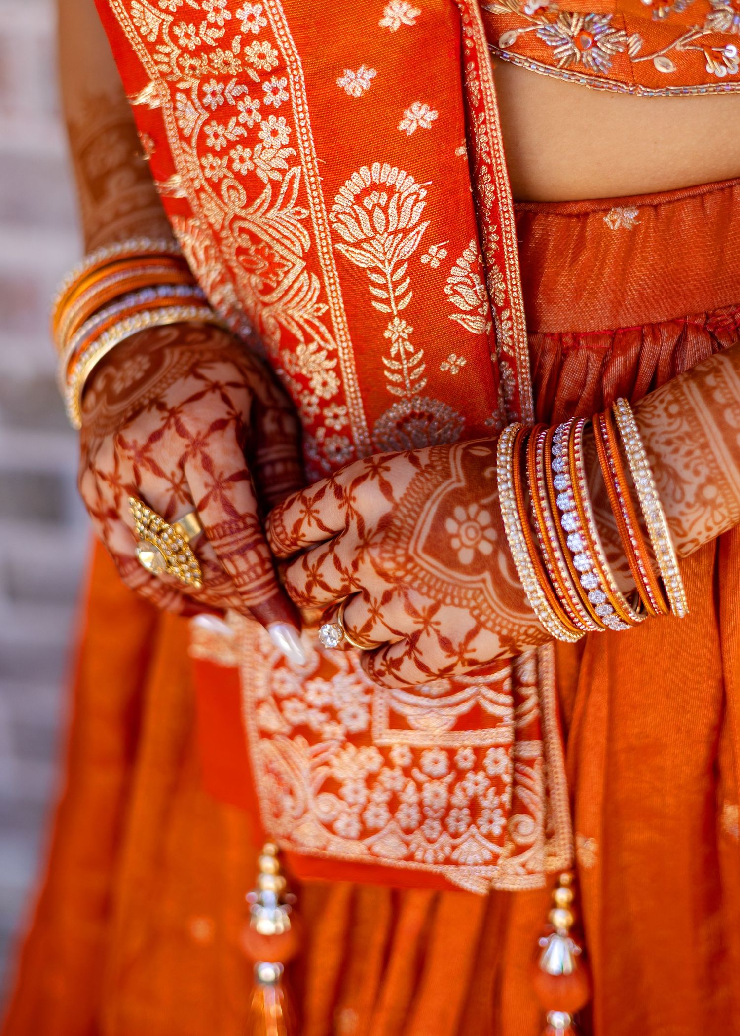 Indian bride in orange lehenga with intricate henna mehndi on hands, gold rings, and colorful bangles.