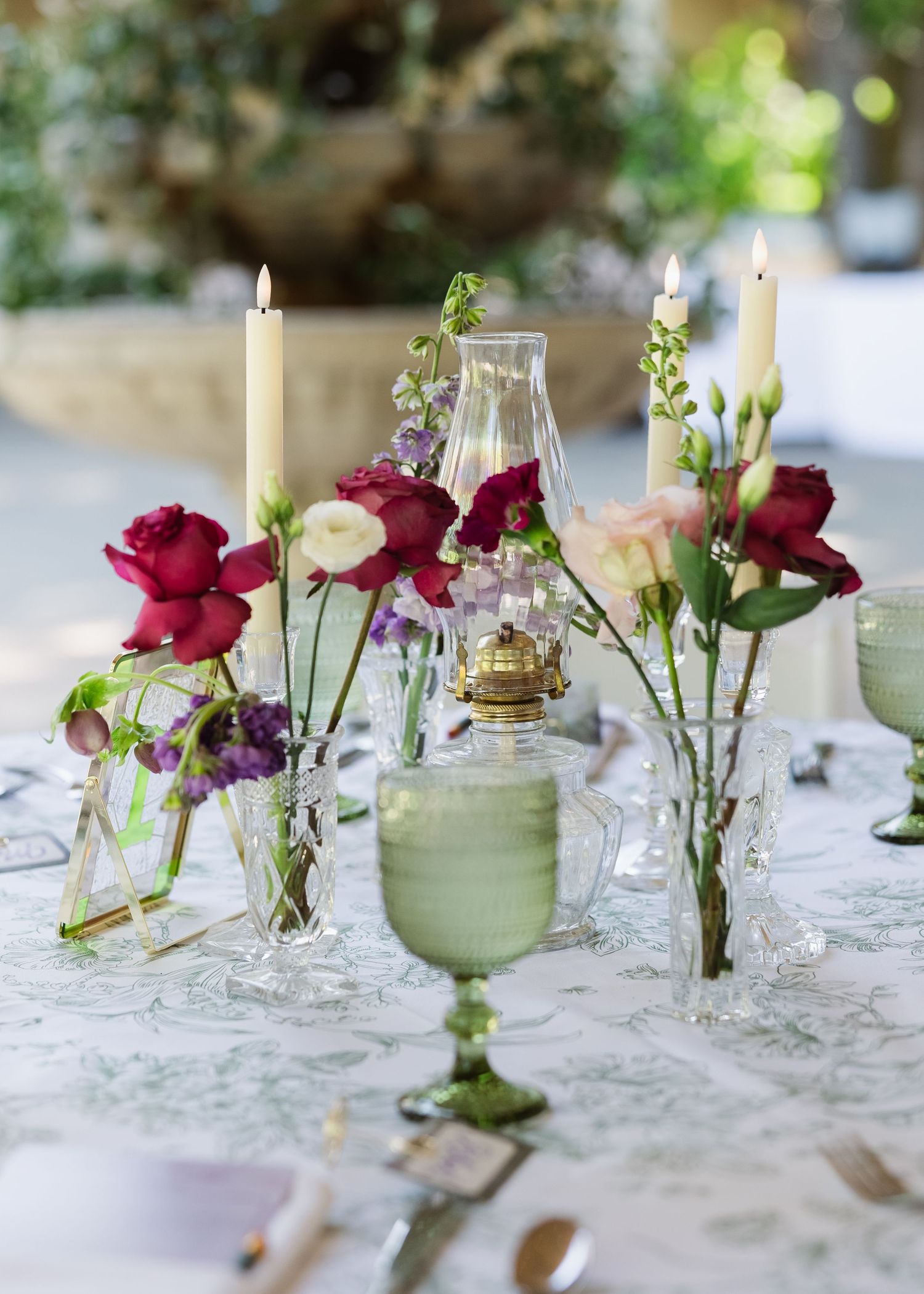 Elegant floral table centerpiece with red roses, candles, and green glassware for a romantic wedding reception.