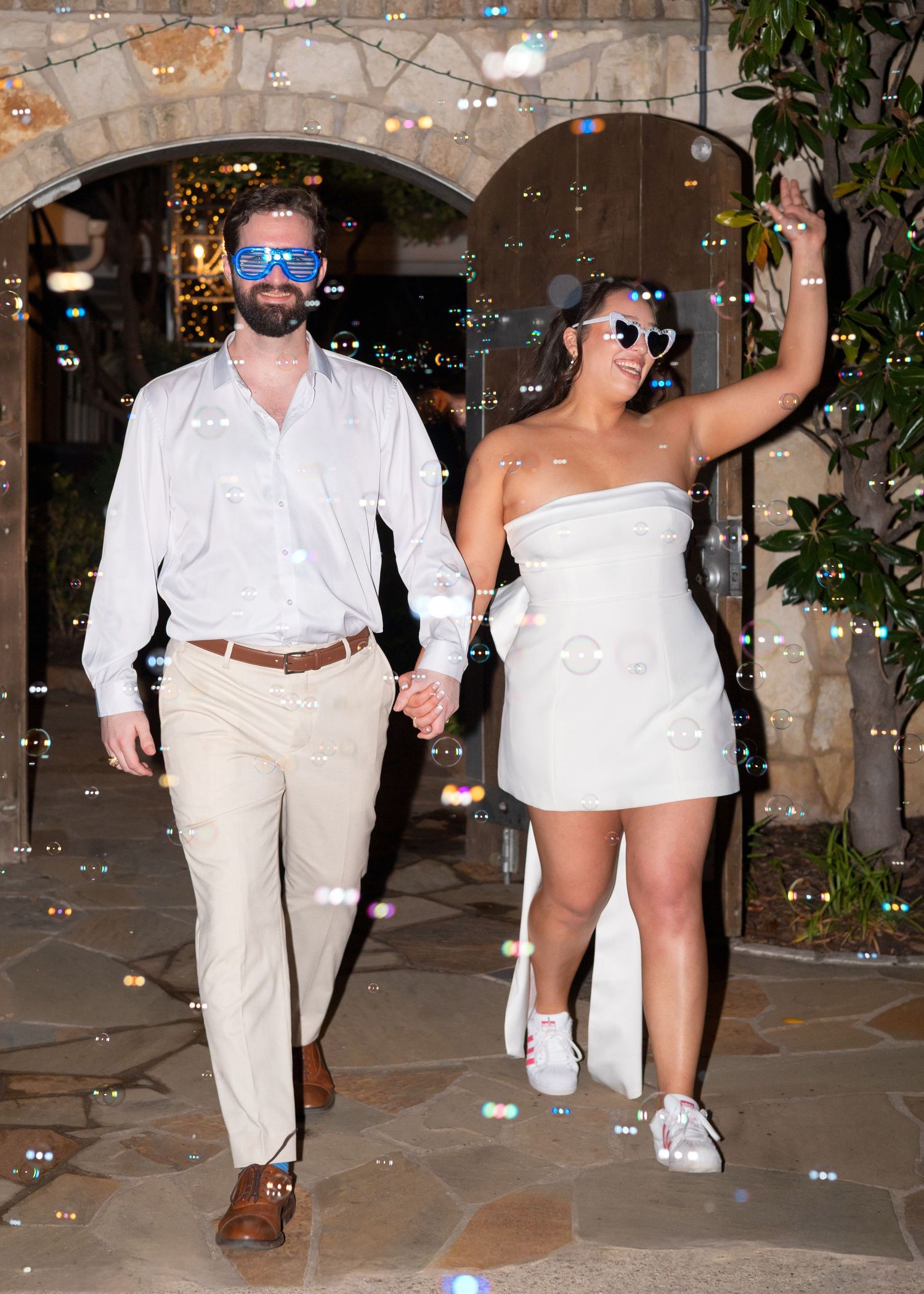 Couple in white outfits with novelty sunglasses walking through bubbles at outdoor wedding reception at night.
