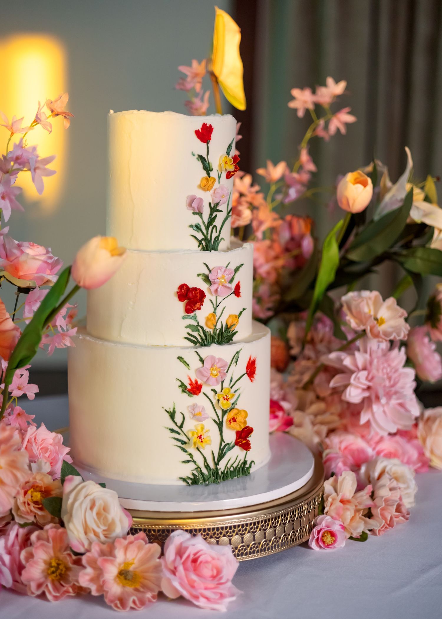 Three-tier white wedding cake with hand-painted wildflowers, displayed on gold stand surrounded by pink roses and tulips.