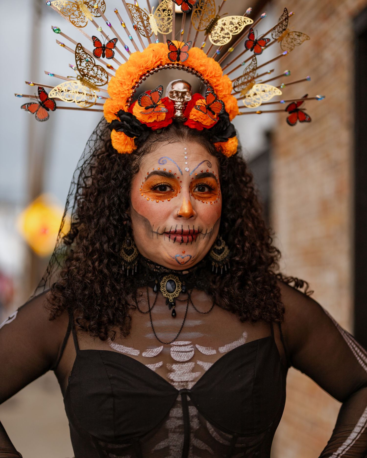 Elaborate Day of the Dead makeup look with orange butterfly crown and dark curly hair.