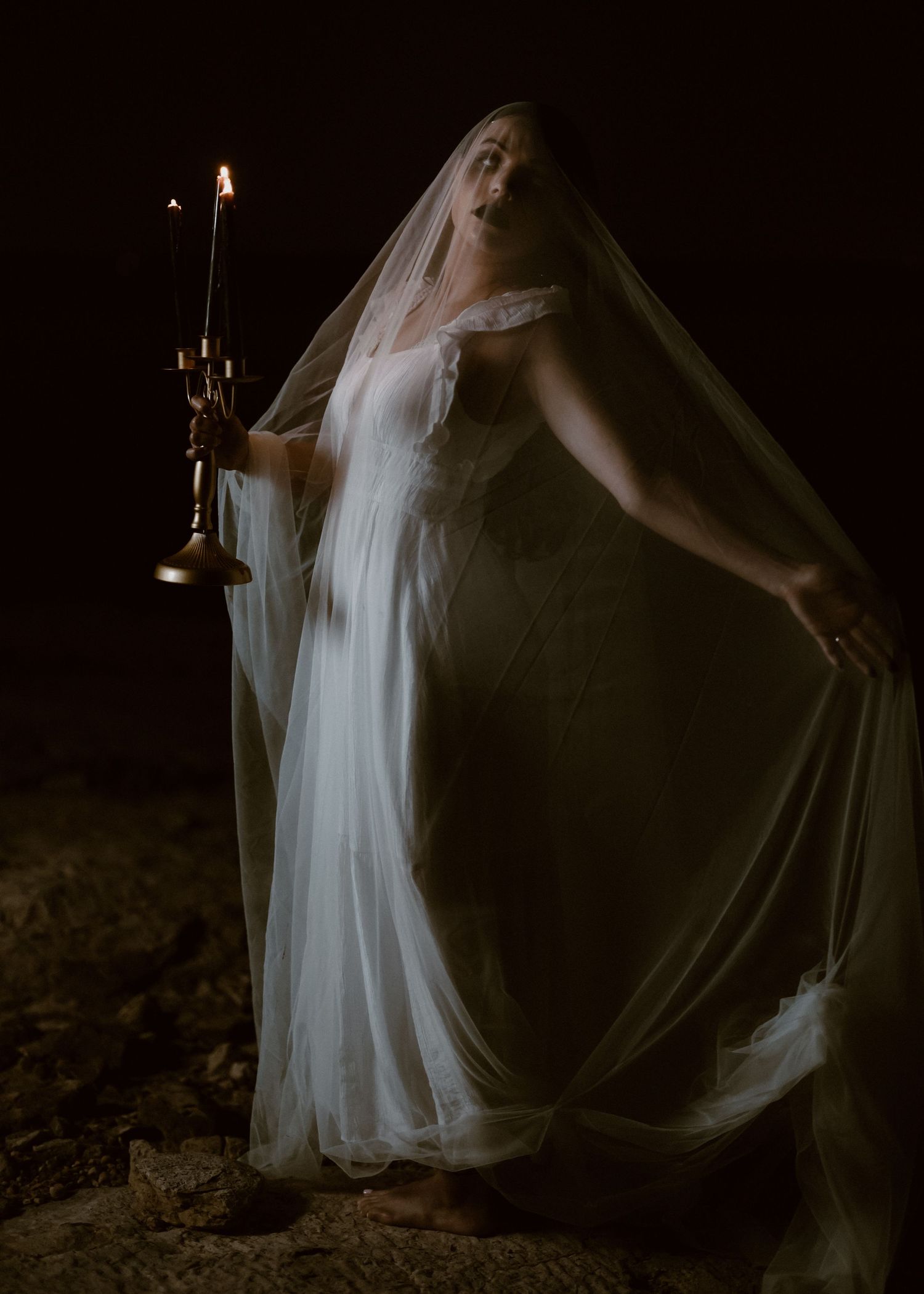 Atmospheric shot of veiled figure in white dress holding candle in dark setting.