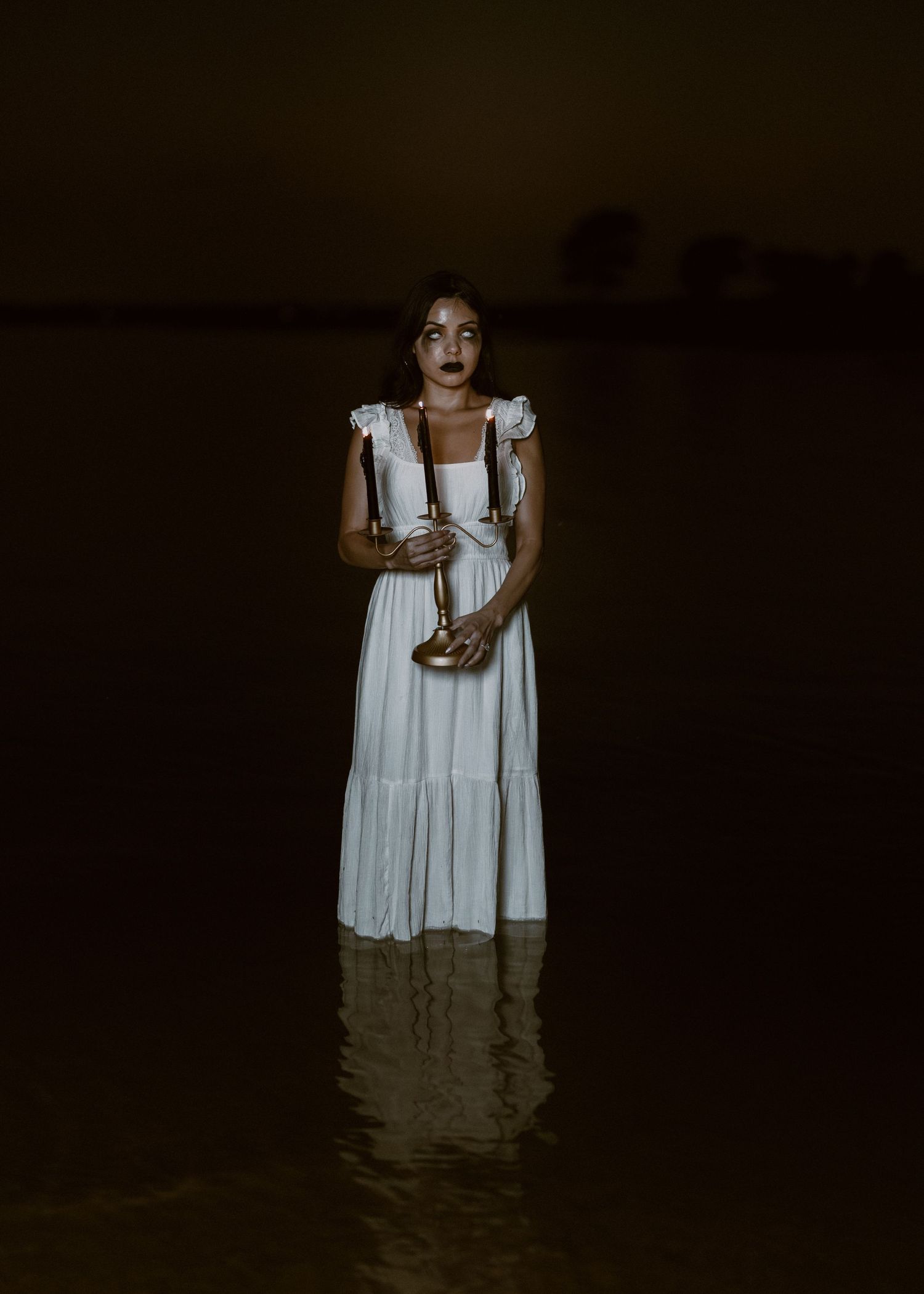 Haunting image of figure in white dress standing in dark water holding candle.