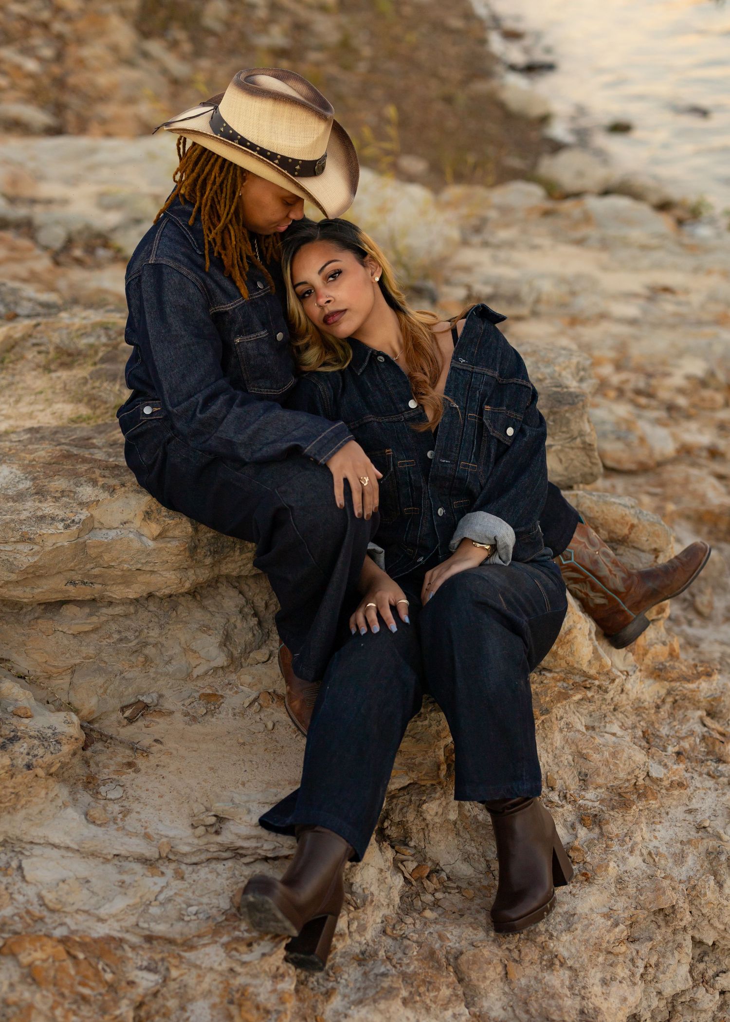 Couple in matching dark outfits and western boots relaxing on rocky outcrop at golden hour.
