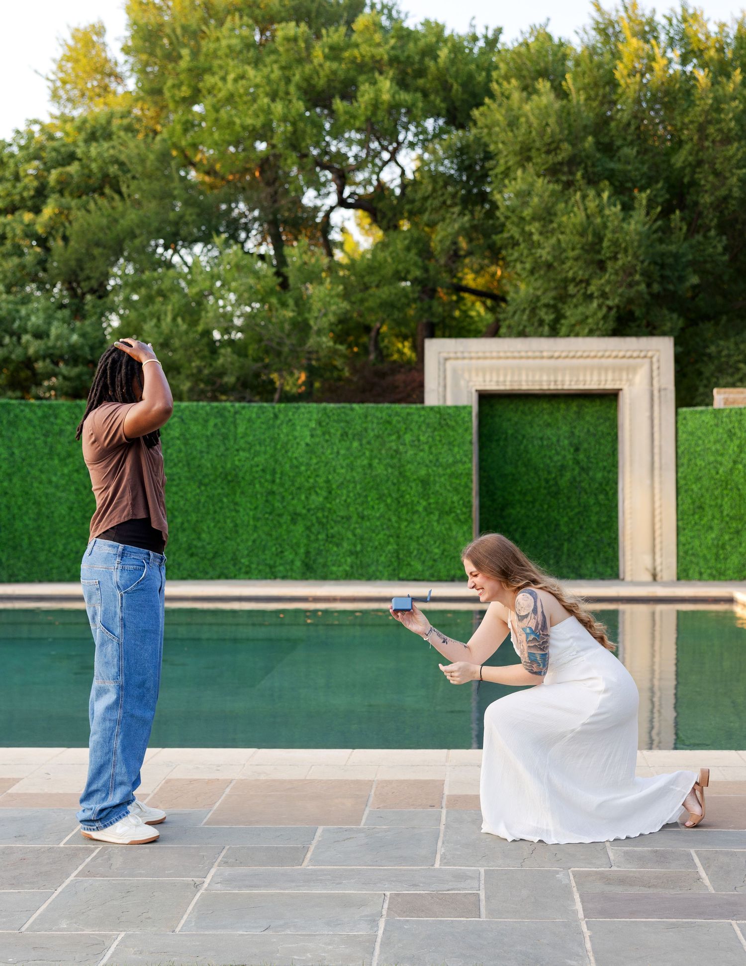 A tattooed woman in a white dress photographs a man posing by a green hedge wall near a pool outdoors.