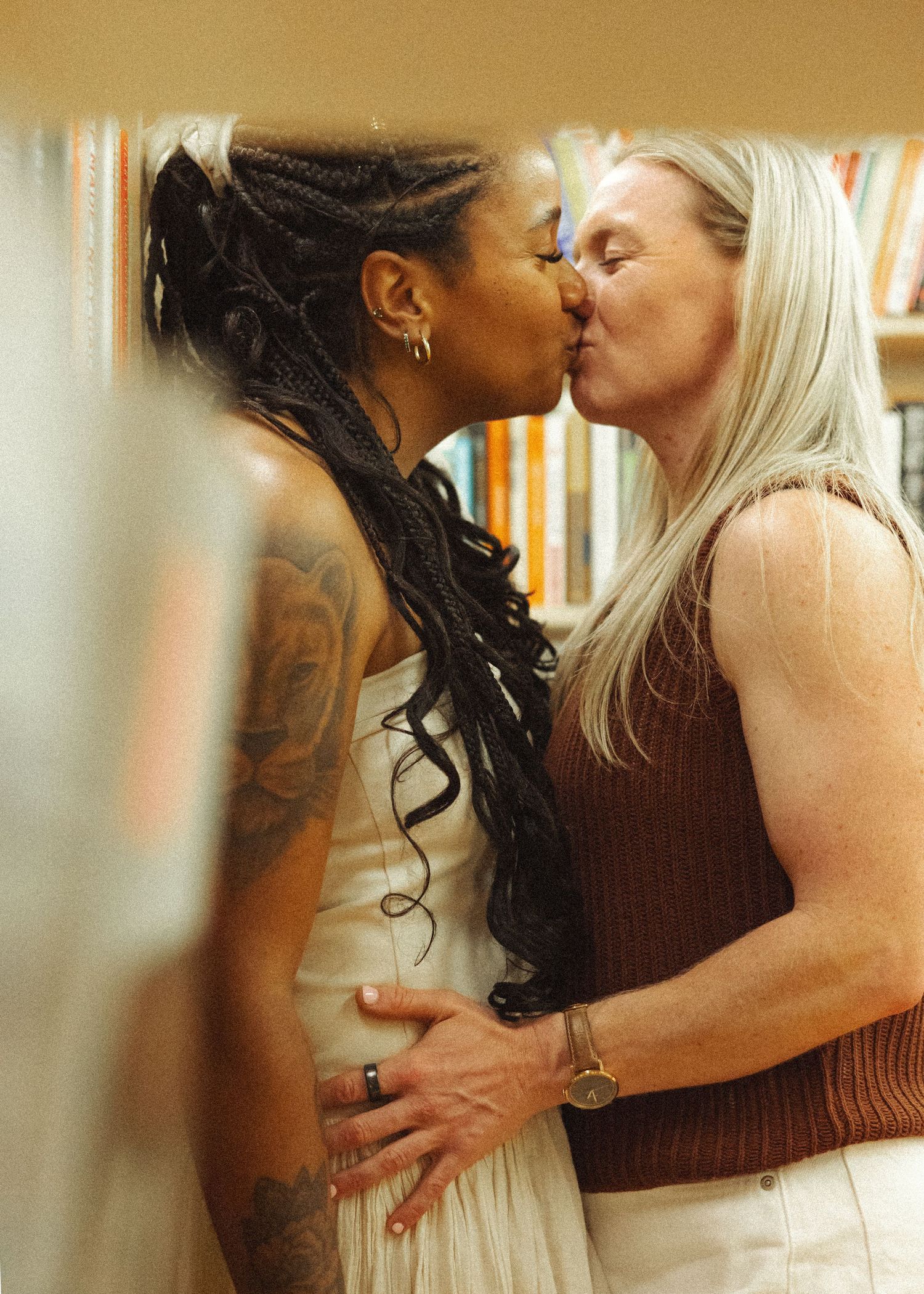 Two women sharing a romantic kiss in a bookstore, one with braided hair and tattoos, the other with long blonde hair.