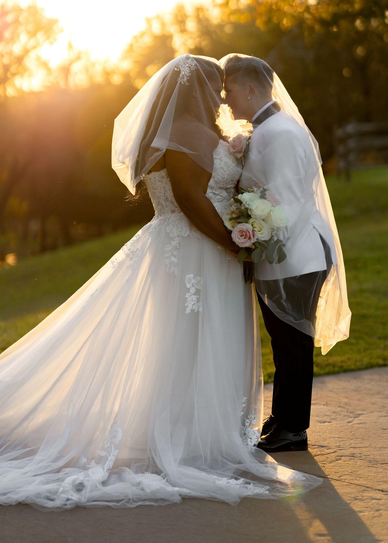 Bride and groom sharing a romantic kiss at golden hour sunset, bride in lace gown with veil holding white bouquet.