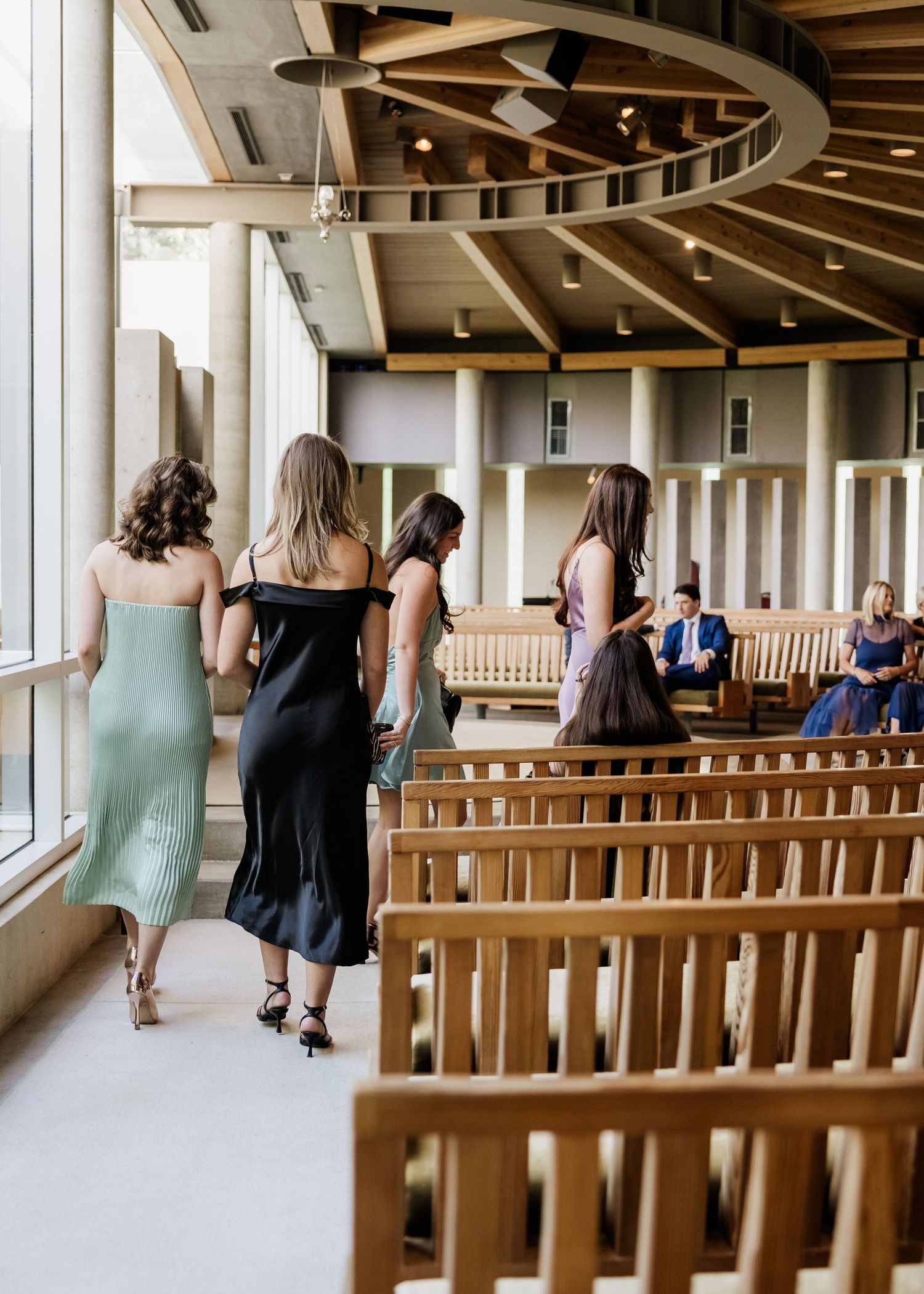 Three women in elegant midi dresses walk down the aisle of a modern wooden chapel during a wedding ceremony.