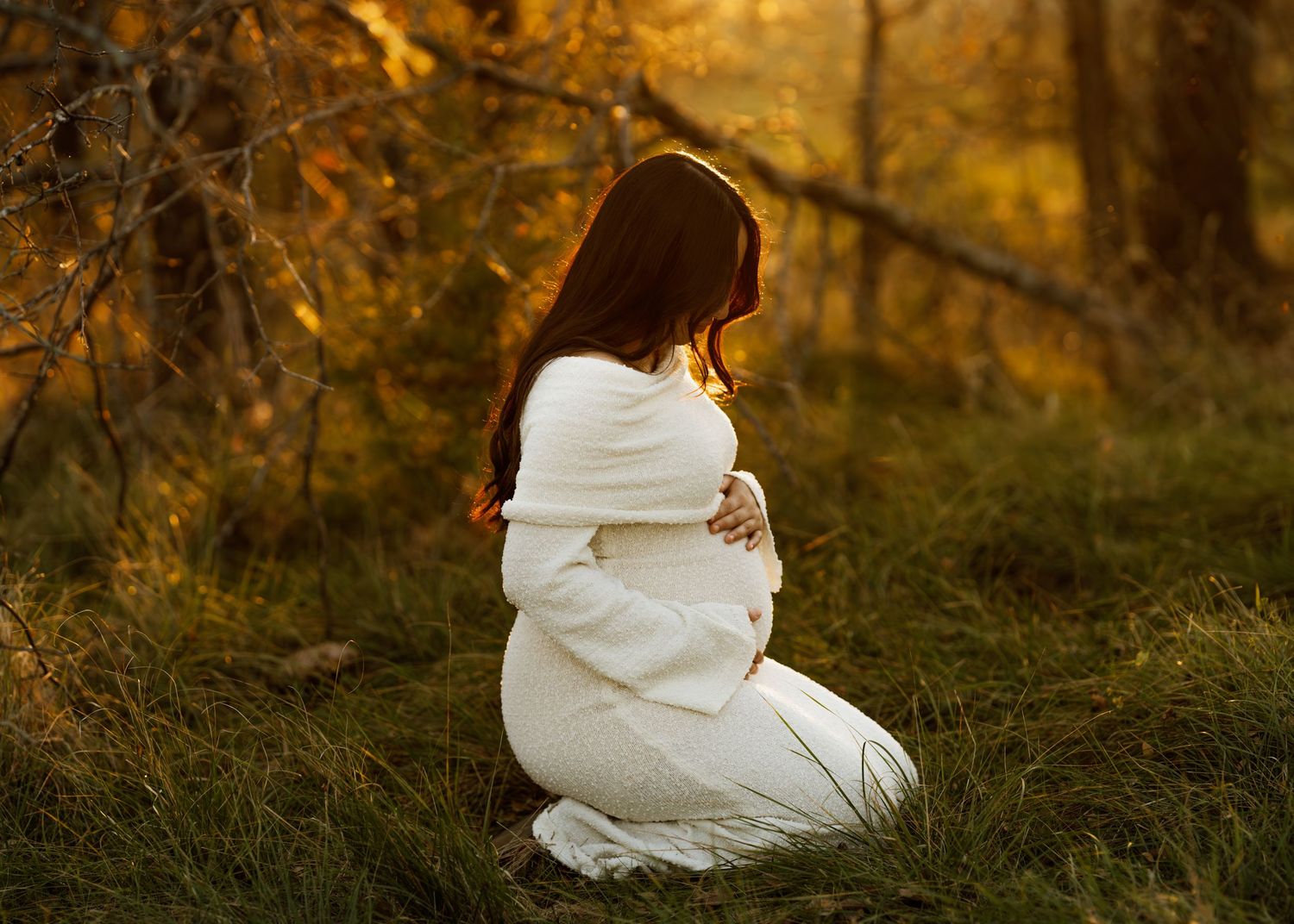 Pregnant woman in white dress kneeling on grass in golden autumn forest light, cradling baby bump.
