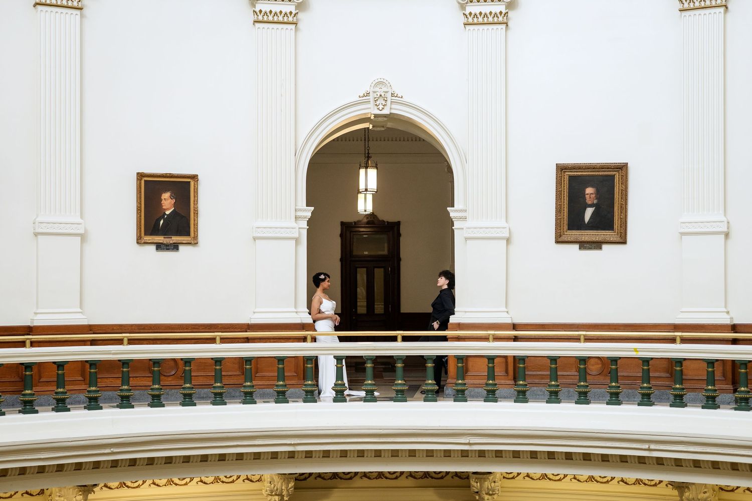 Wedding couple stands together on an ornate balcony in a classical white building with arched doorways and portraits.