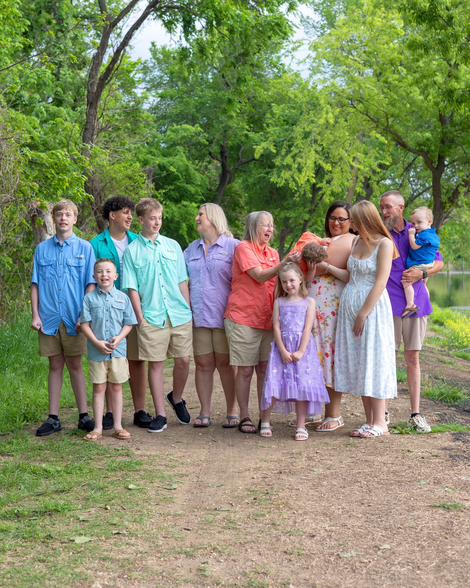 Large family group photo outdoors on a dirt path surrounded by lush green trees, wearing coordinated pastel outfits.