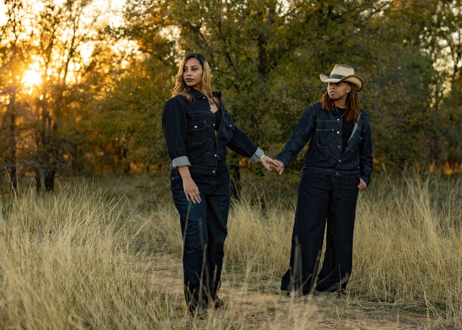 Couple walking through tall grass field during golden sunset light wearing dark clothing.