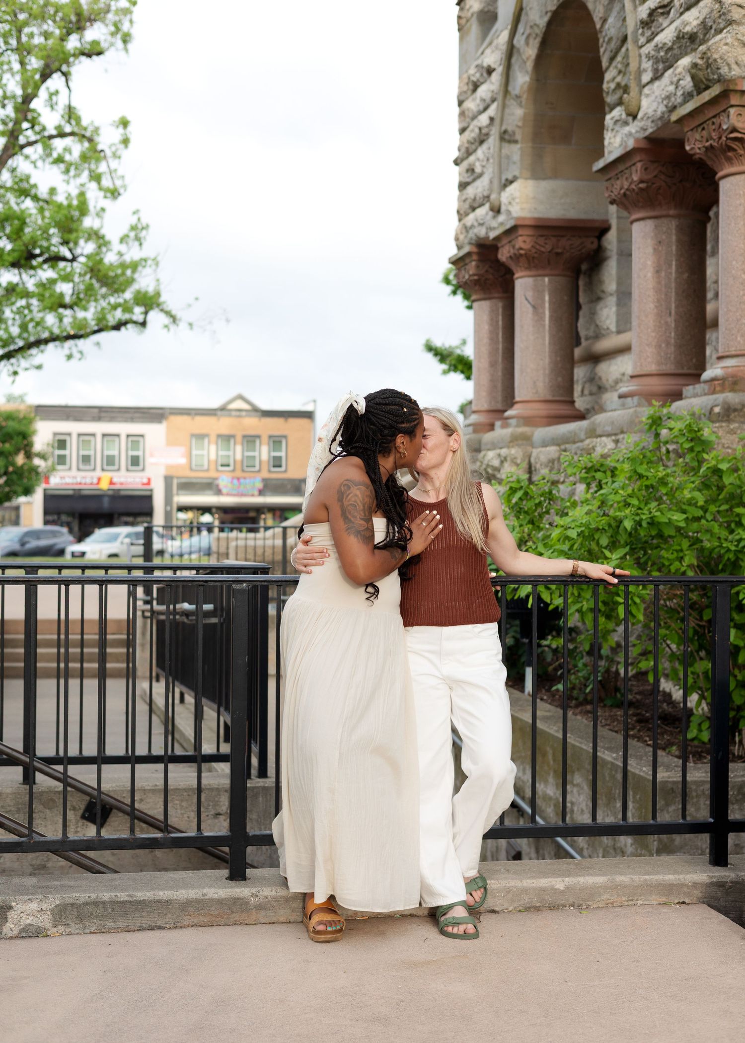 Two women sharing a kiss outside a historic stone building, one in a white dress and one in brown top and white pants.