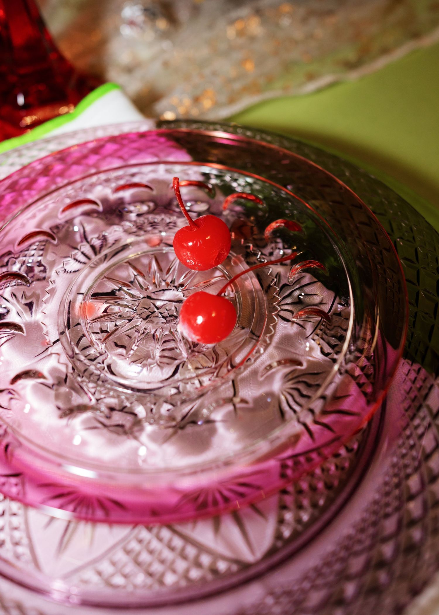 Two maraschino cherries on a decorative cut crystal glass dish with pink accents on a green surface.