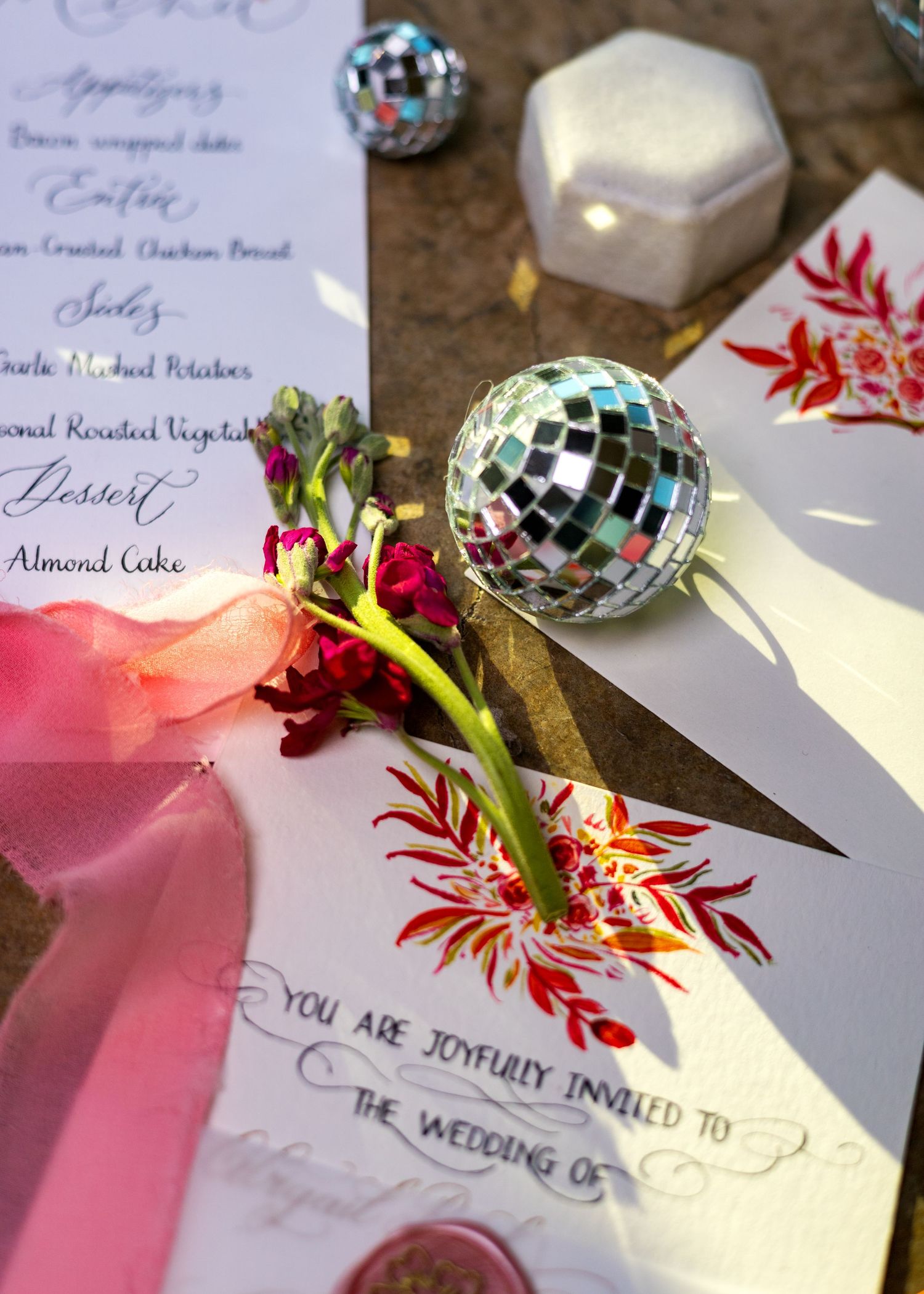 Wedding invitation with mini disco ball, red floral details, and menu card styled on a gold table.